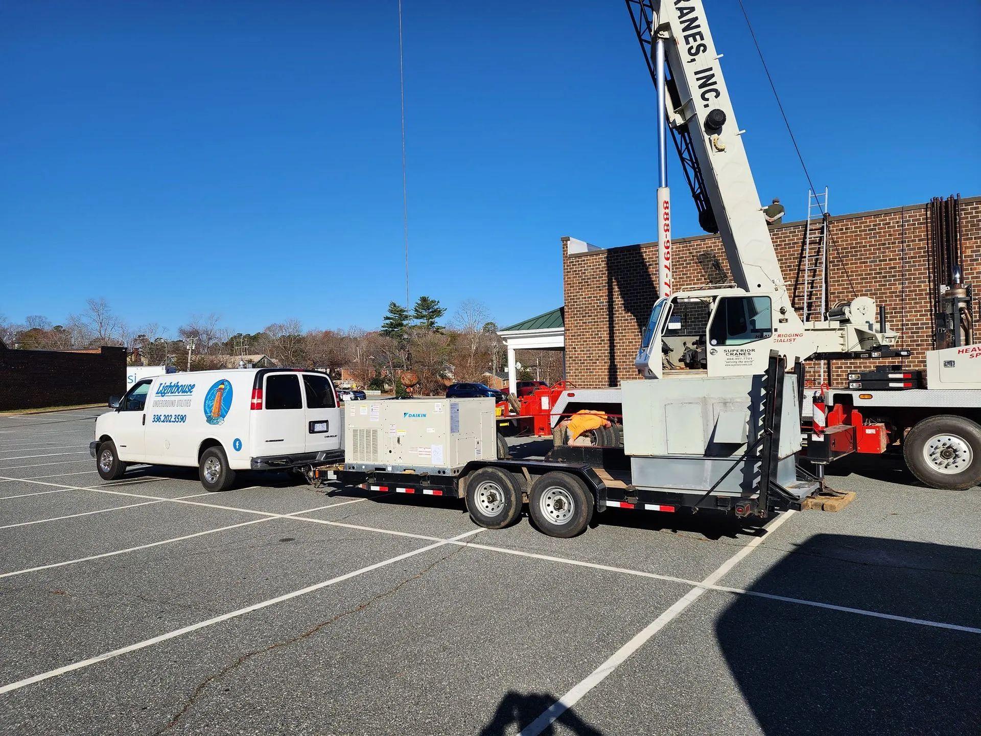 Utility truck and trailer with raised crane parked in a lot beside a brick building