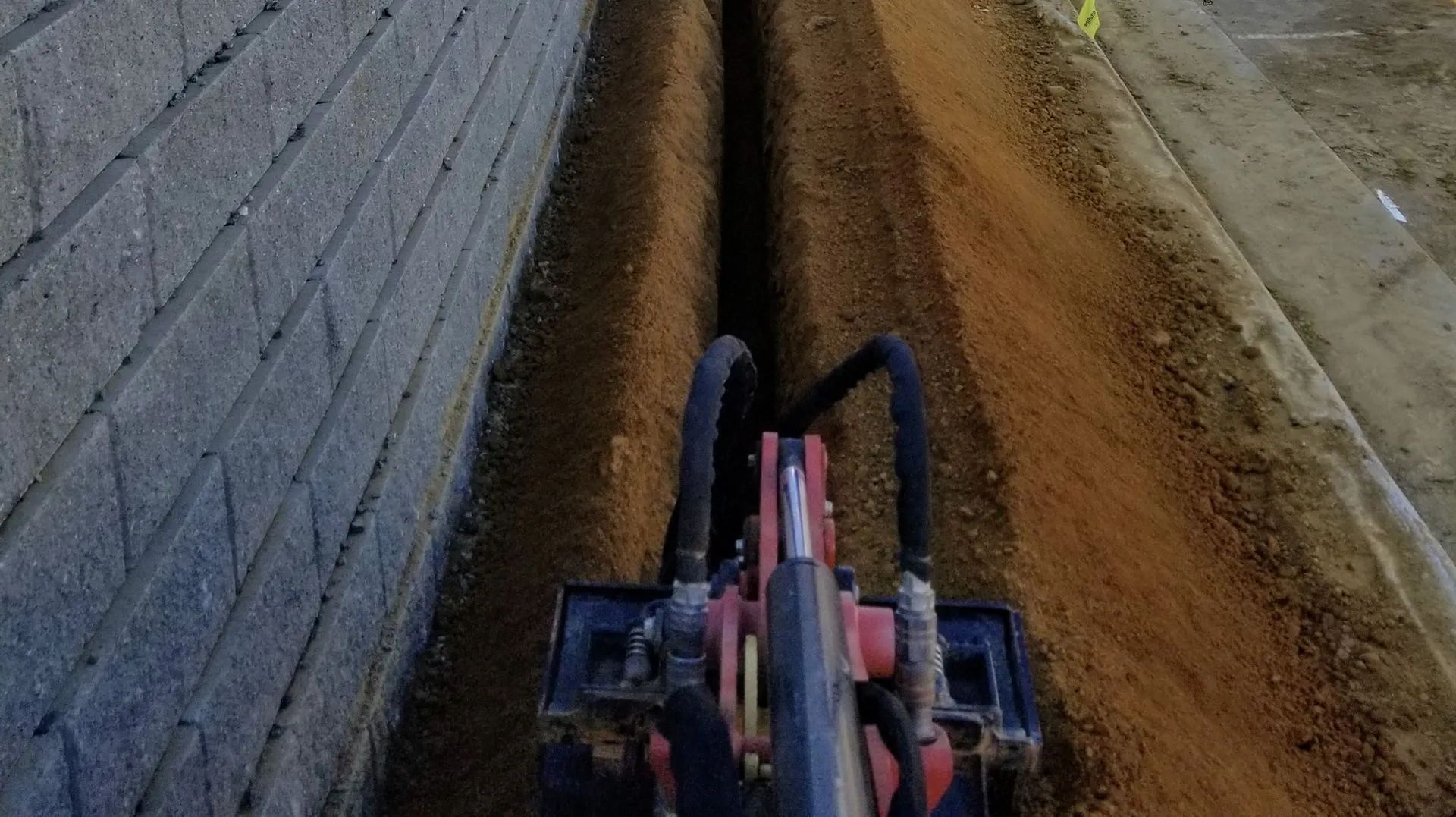 Forward view from a tractor in a narrow dirt trench beside a brick wall.