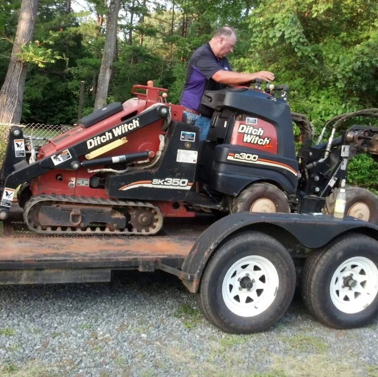 Man on a trailer drives a red Ditch Witch track loader in a wooded outdoor area.