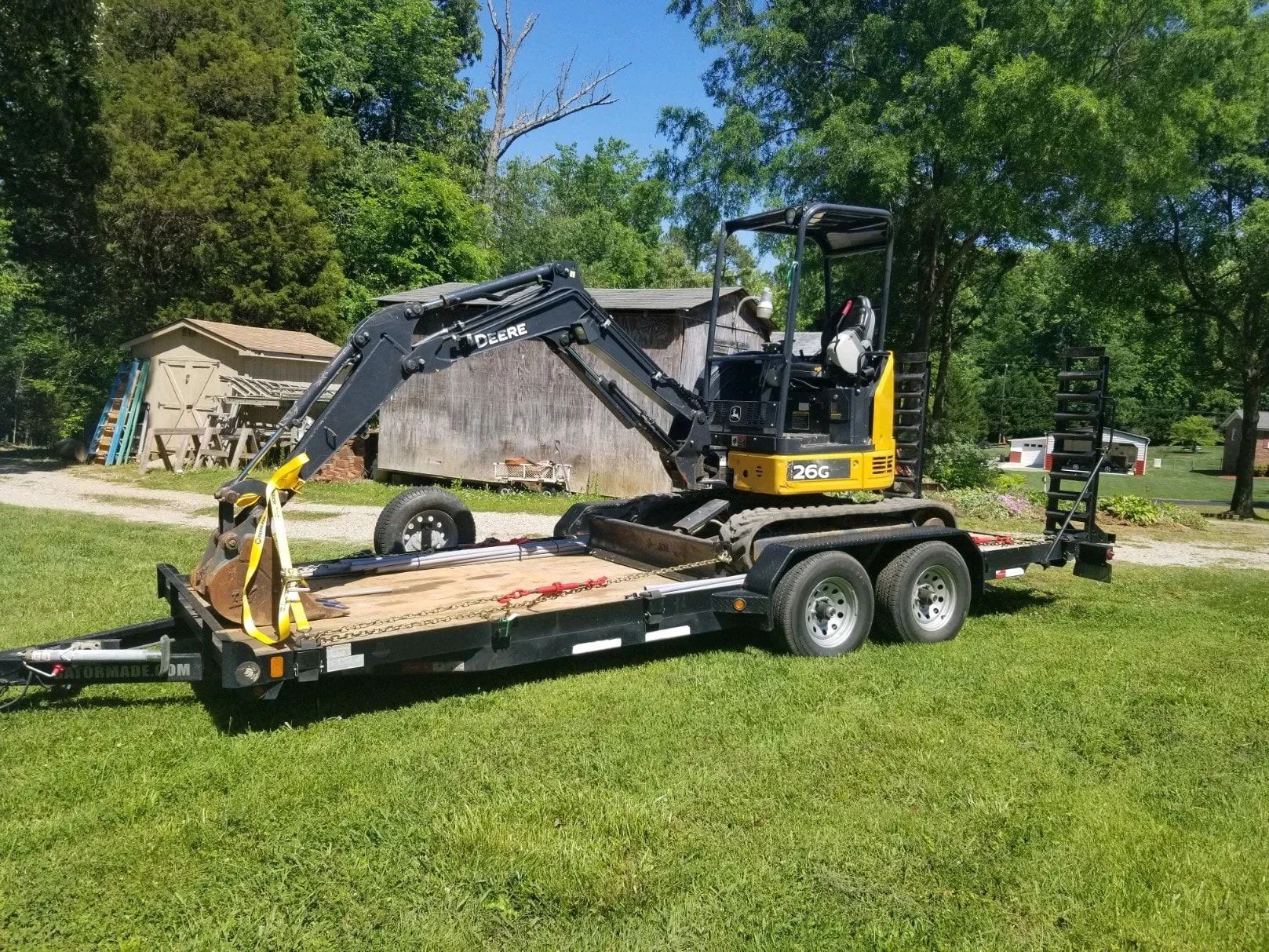Compact excavator on a trailer in a grassy yard beside a shed, with trees and a blue sky.