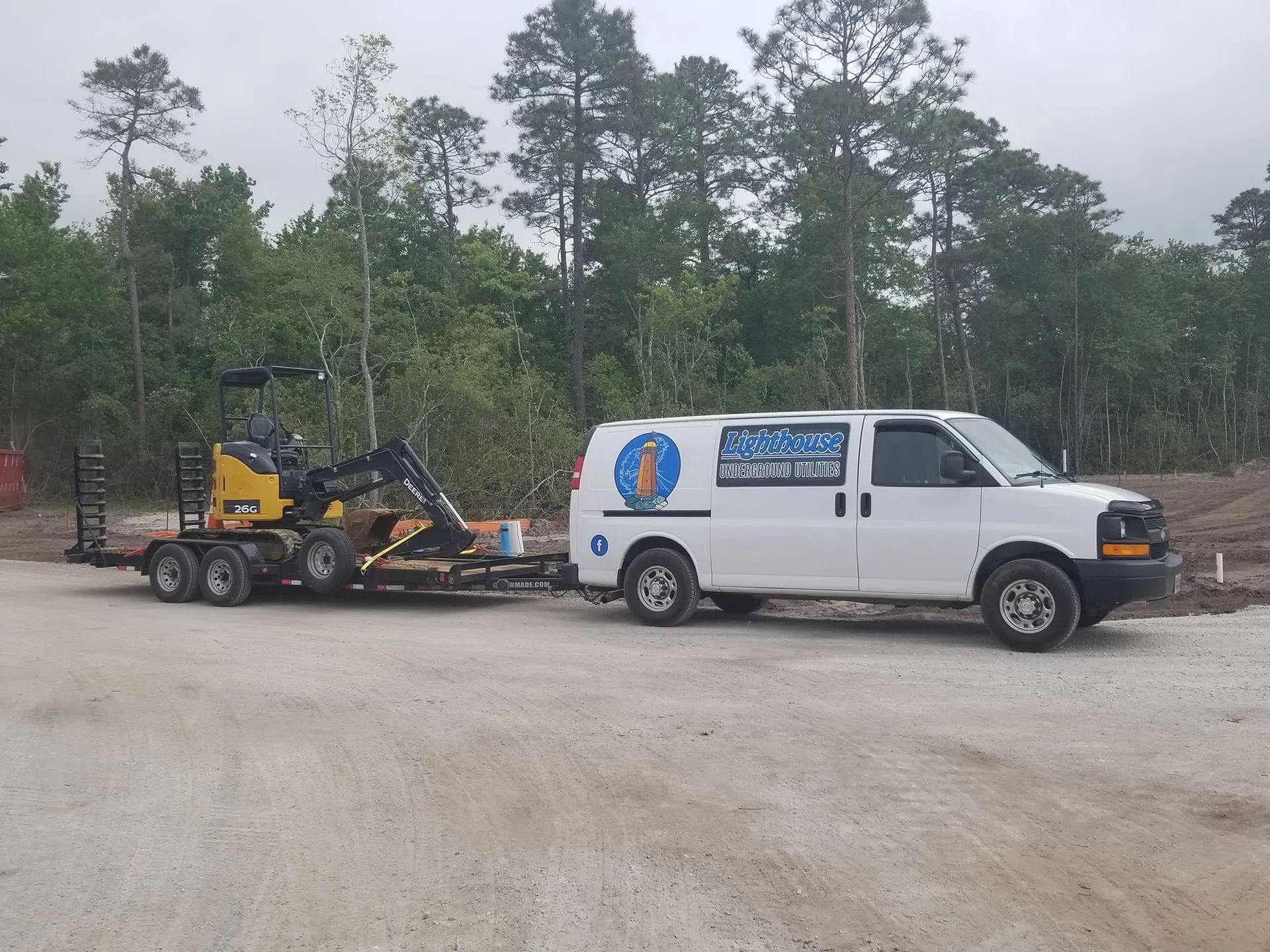 White service van towing a yellow skid steer on a flatbed trailer in a wooded gravel lot.
