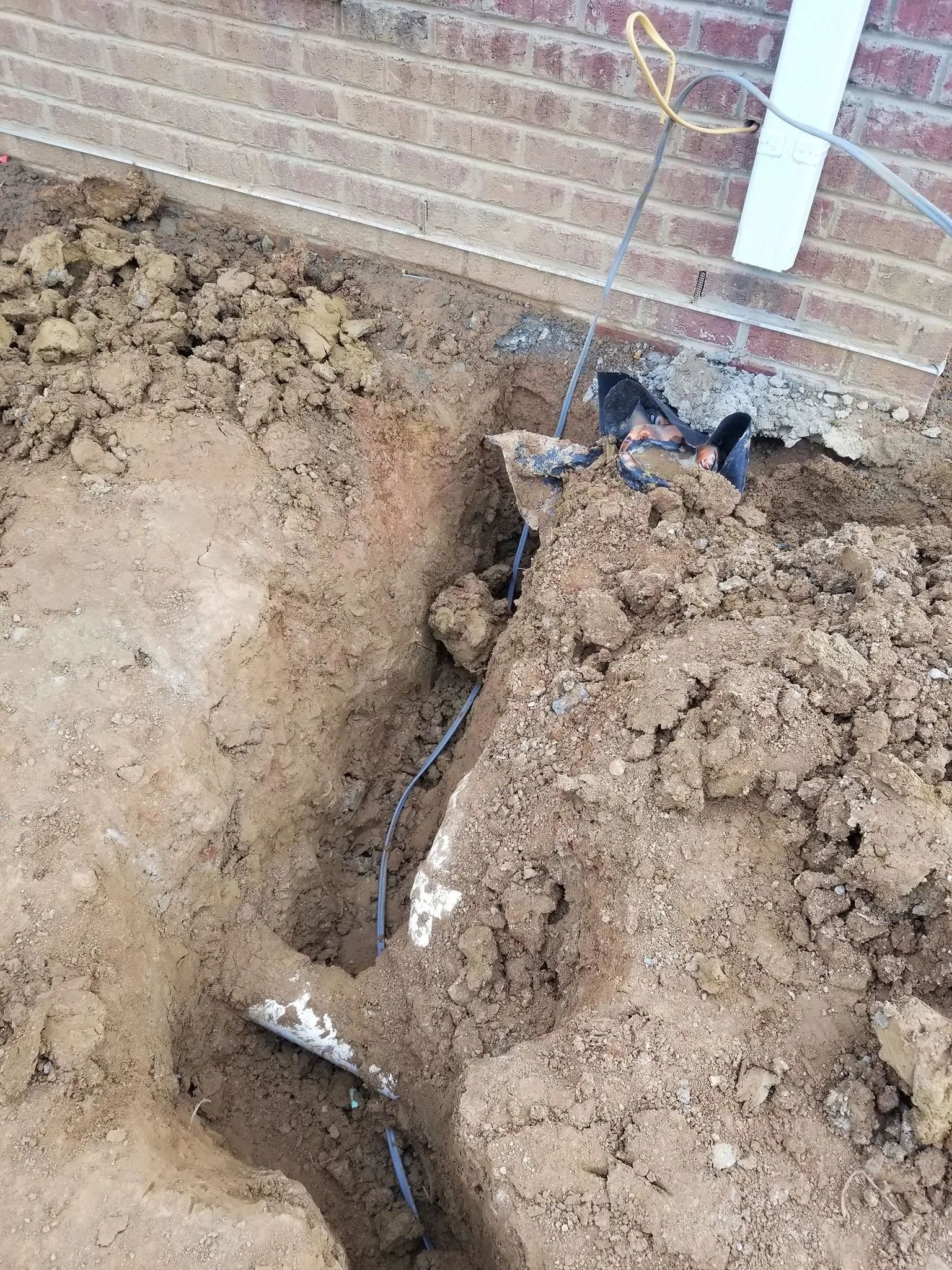Open trench beside a brick wall with exposed black utility cables in loose dirt