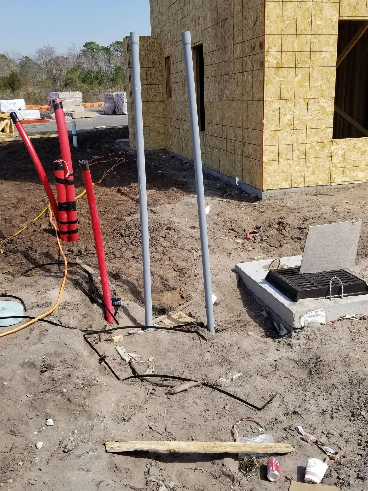 Construction site with foundation walls, red safety barrier, and exposed utility pipes in dirt ground.