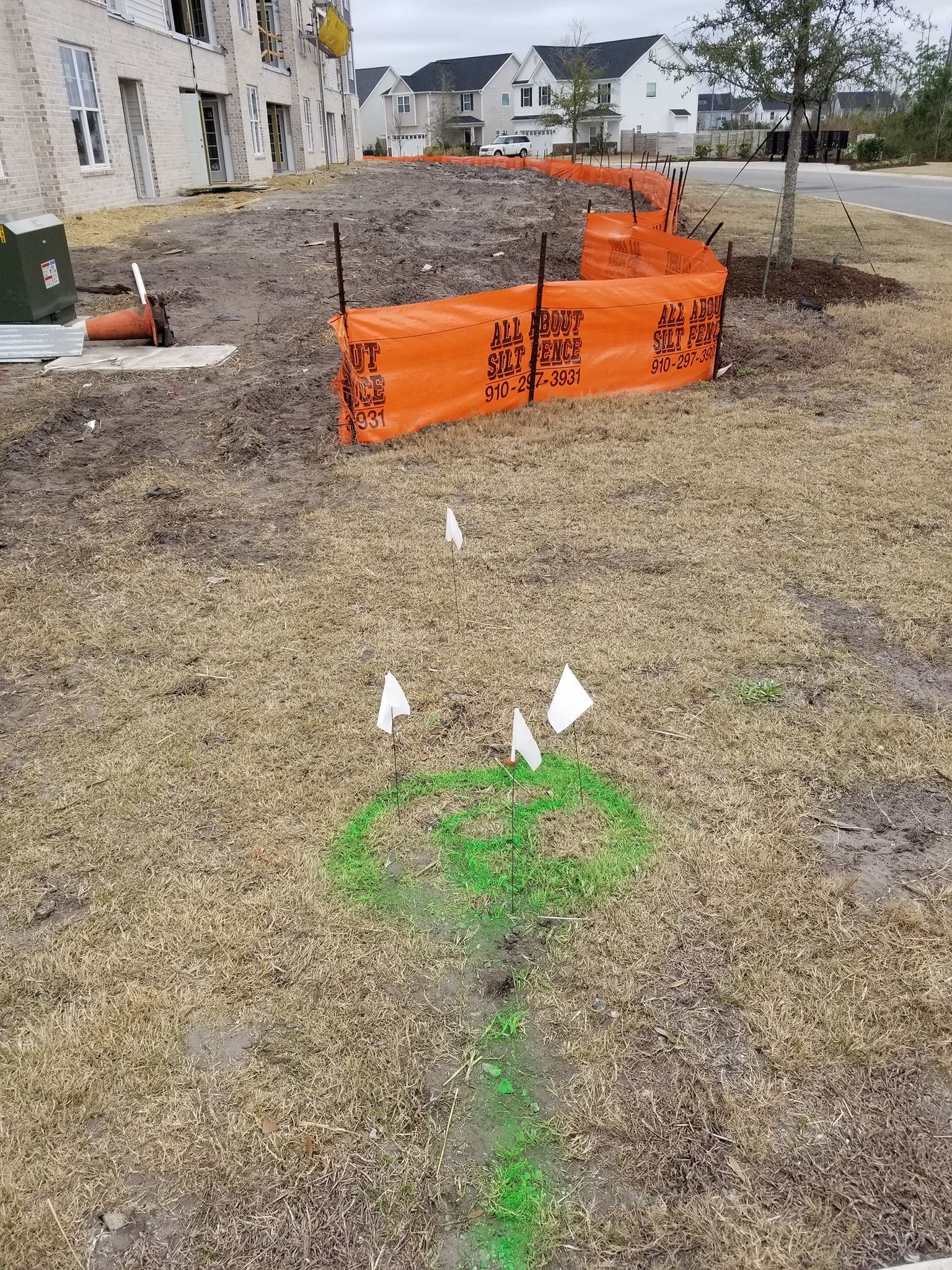 Orange construction barrier in a grassy yard, with green spray markings and white flags marking utility work.