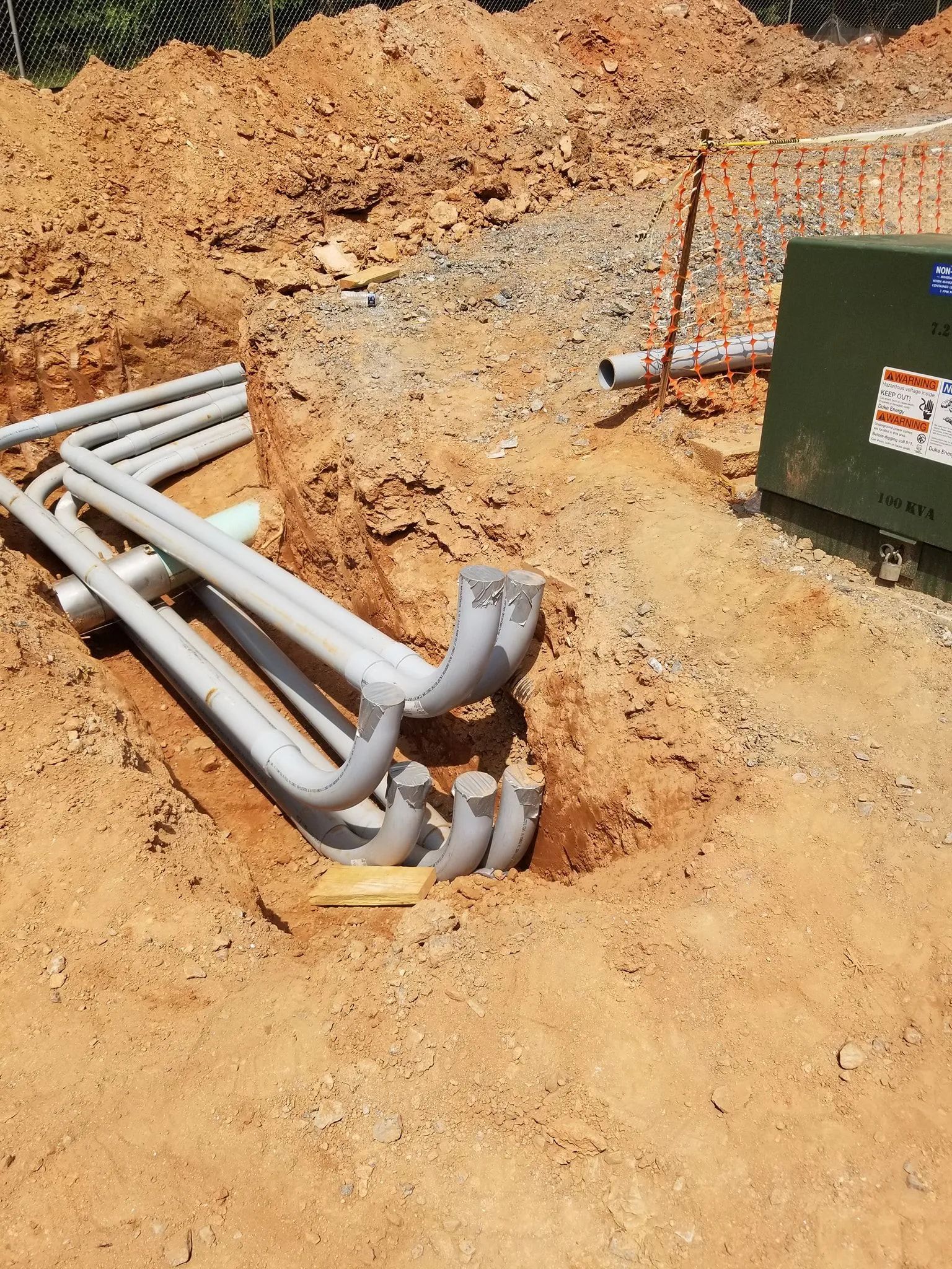 Trenched gray utility pipes and conduit on dirt beside a green electrical box at a construction site