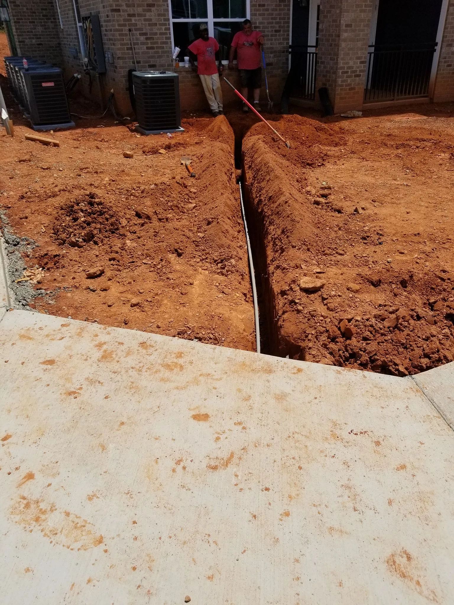 Deep trench cut through red dirt beside a concrete slab and building, with workers in the distance