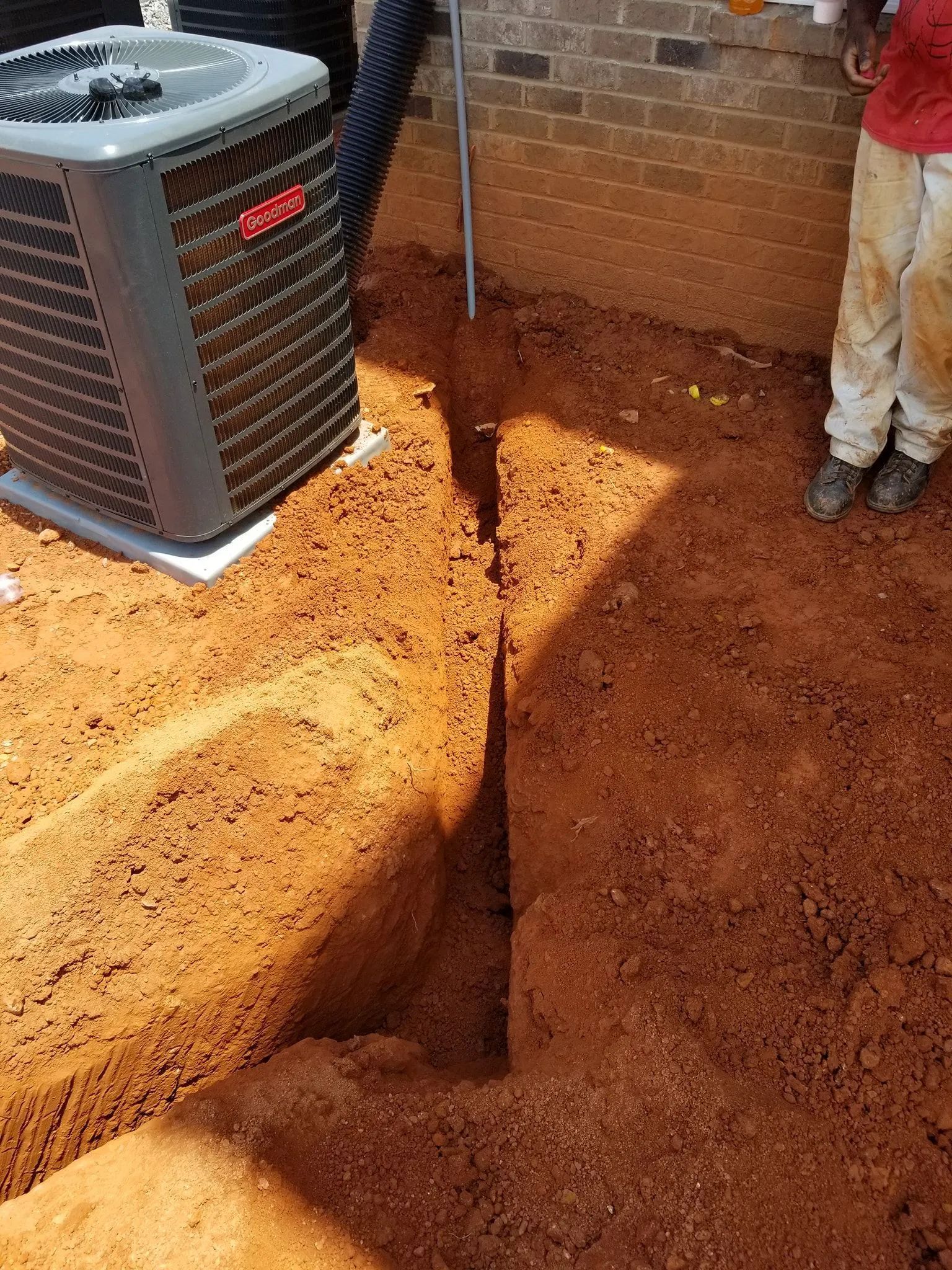 Deep trench beside an outdoor HVAC unit near a brick wall, with a worker standing nearby.