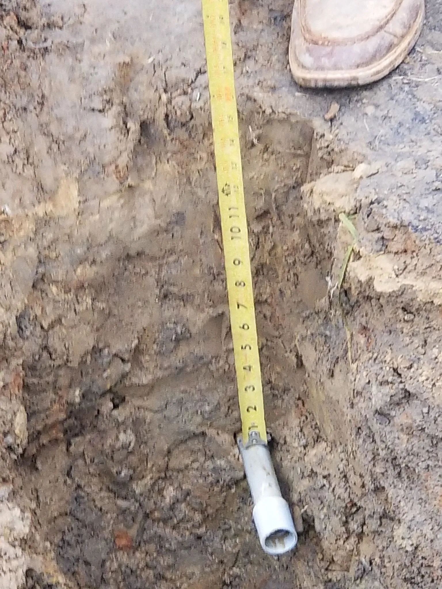 Yellow measuring tape in a dirt trench beside a white pipe, with a boot visible at the top.
