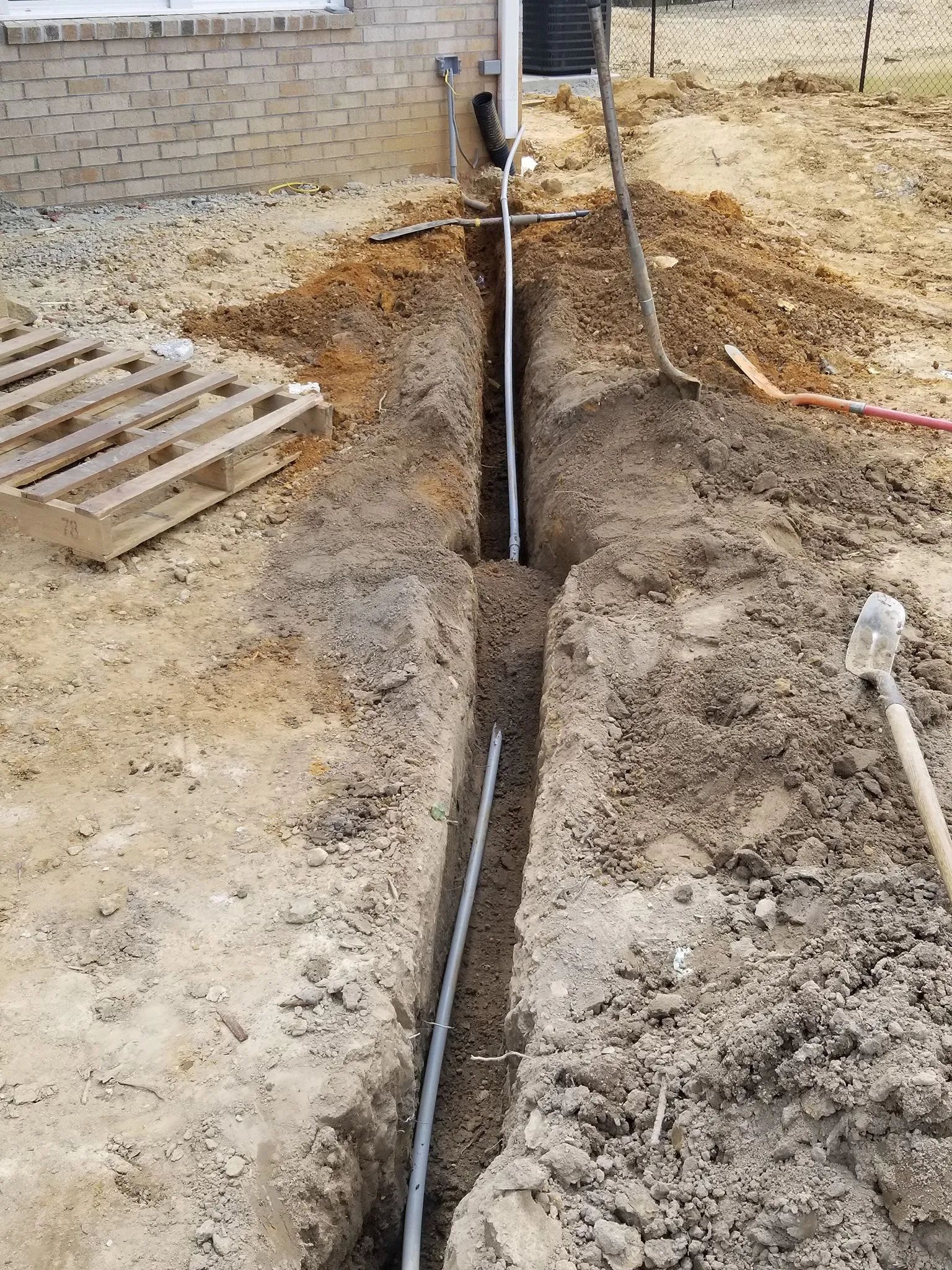 Trench dug along a construction site, with exposed pipes and dirt piles beside a brick wall.