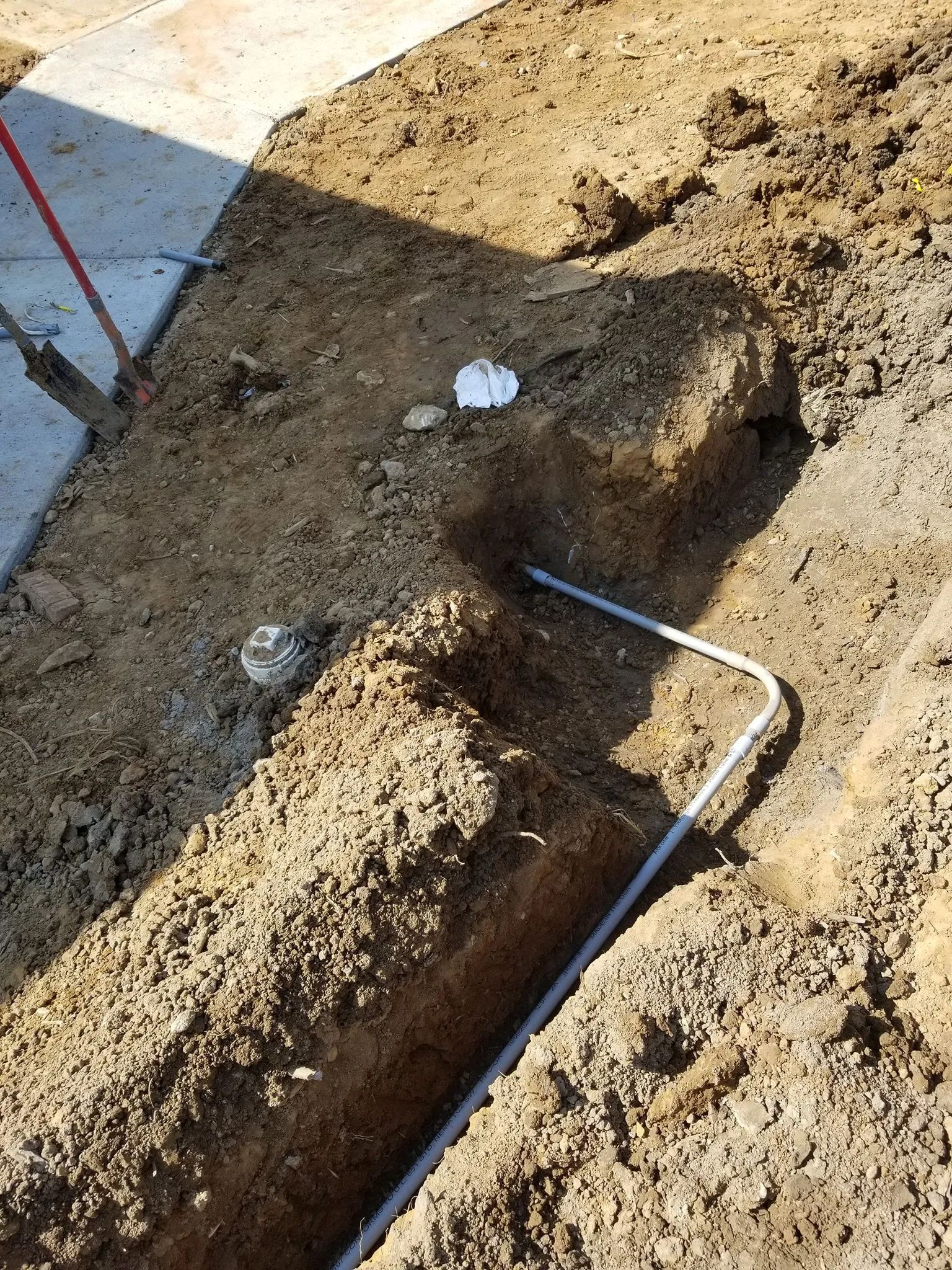 Open trench in dirt beside a sidewalk, with a bent metal pipe exposed in the ground.