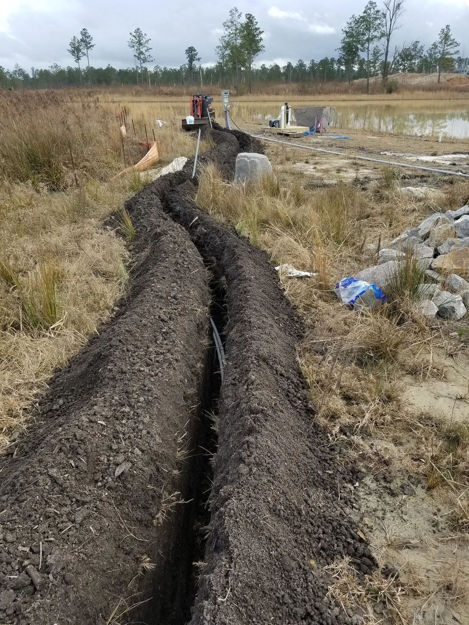 Long dark trench cut through dry grass in a rural field, with workers and equipment in the distance.