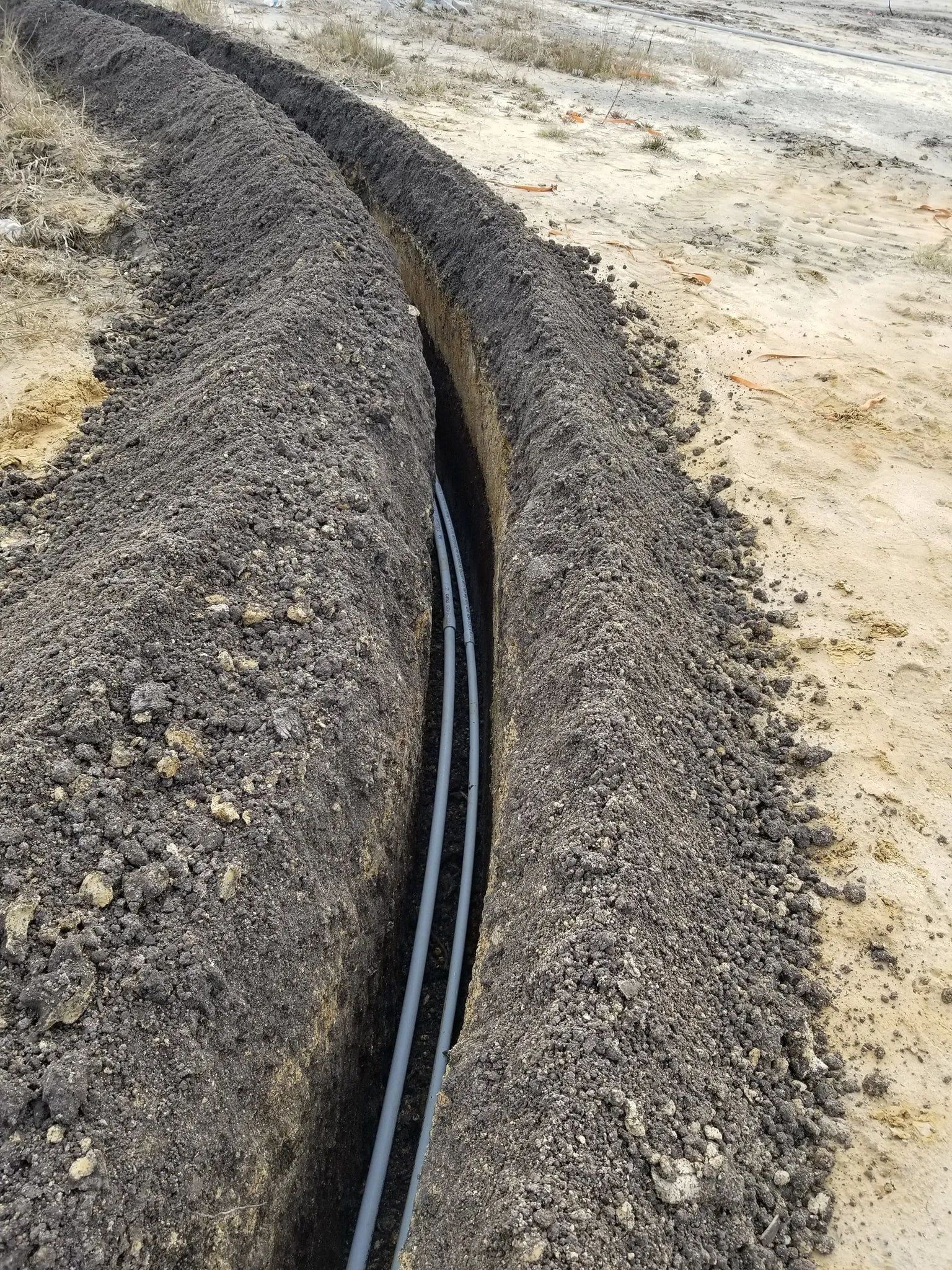 Black utility cables laid in a narrow trench through sandy ground, lined with dark soil.