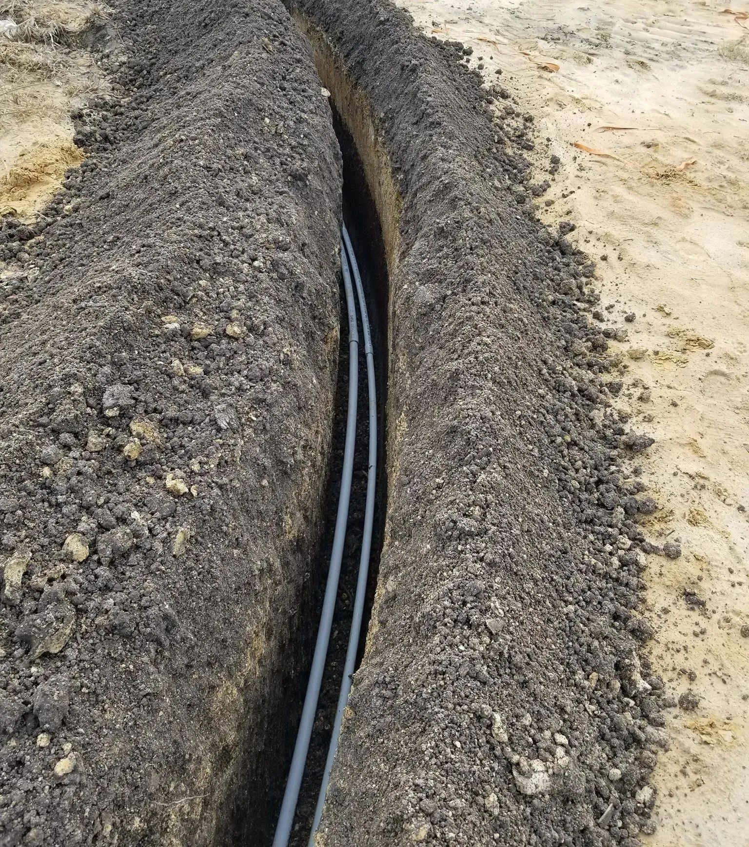 A narrow trench with black cables running through gravel along a sandy construction site
