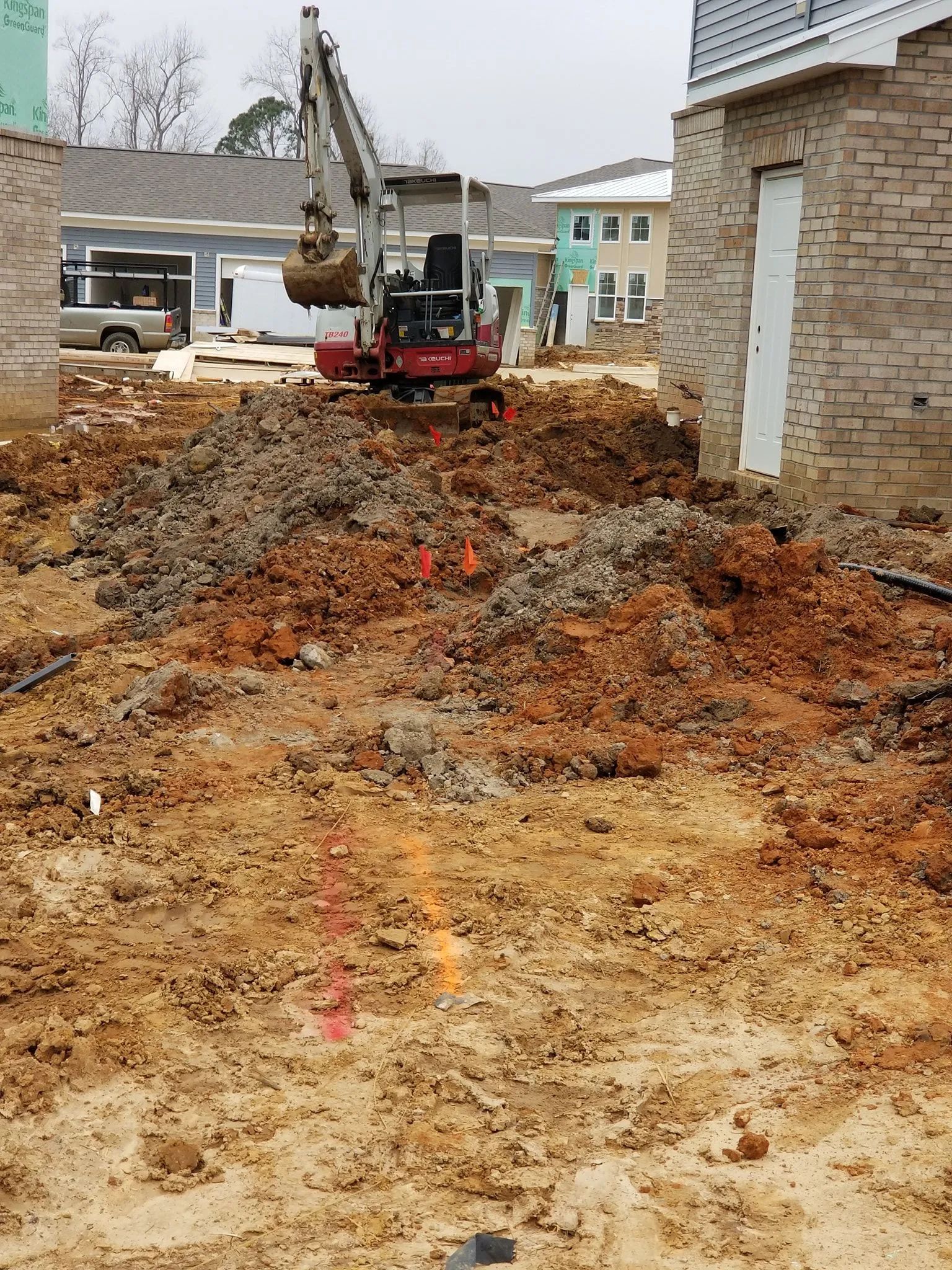 Excavator digging a muddy trench between houses at a construction site