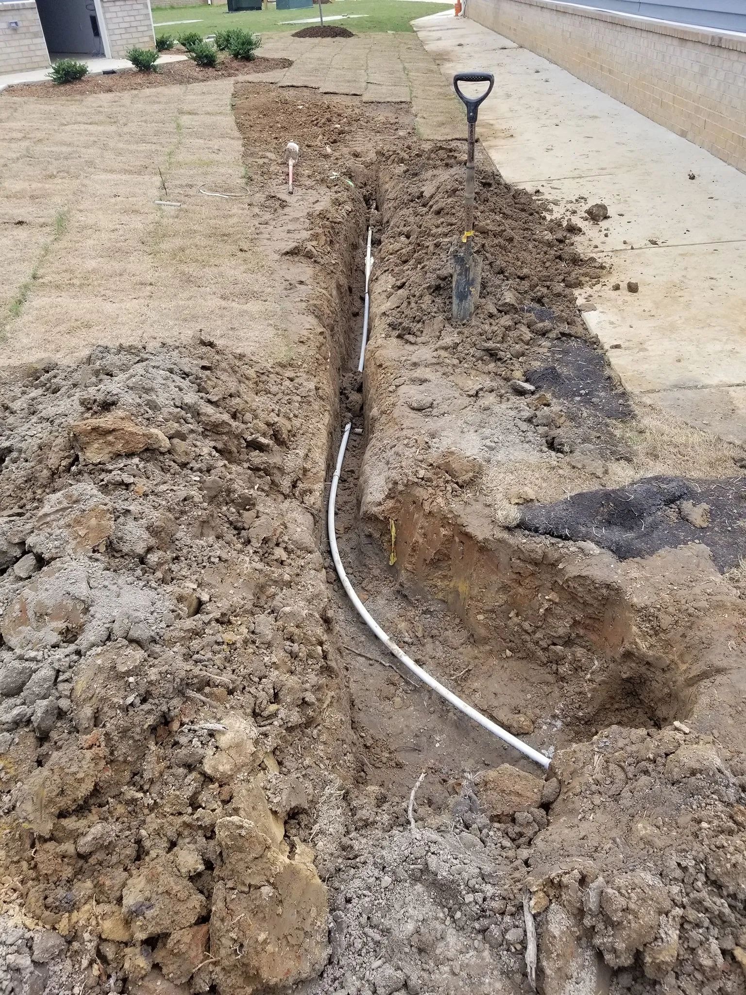 Trench dug beside a sidewalk with a white pipe exposed in the dirt, near a small solar path light.