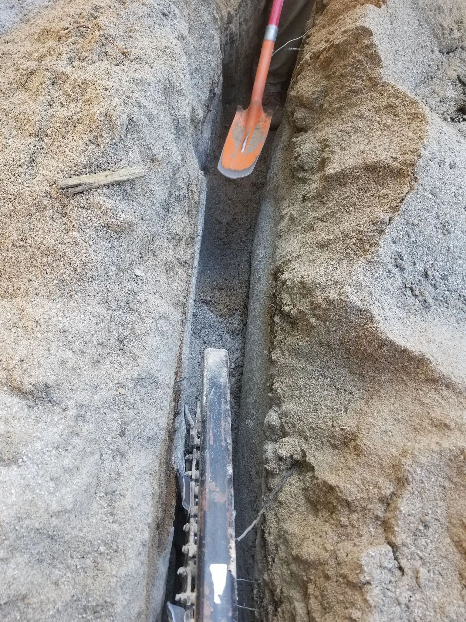 Narrow trench between rocky ground, with a tool handle and a black cable or pipe running down the center