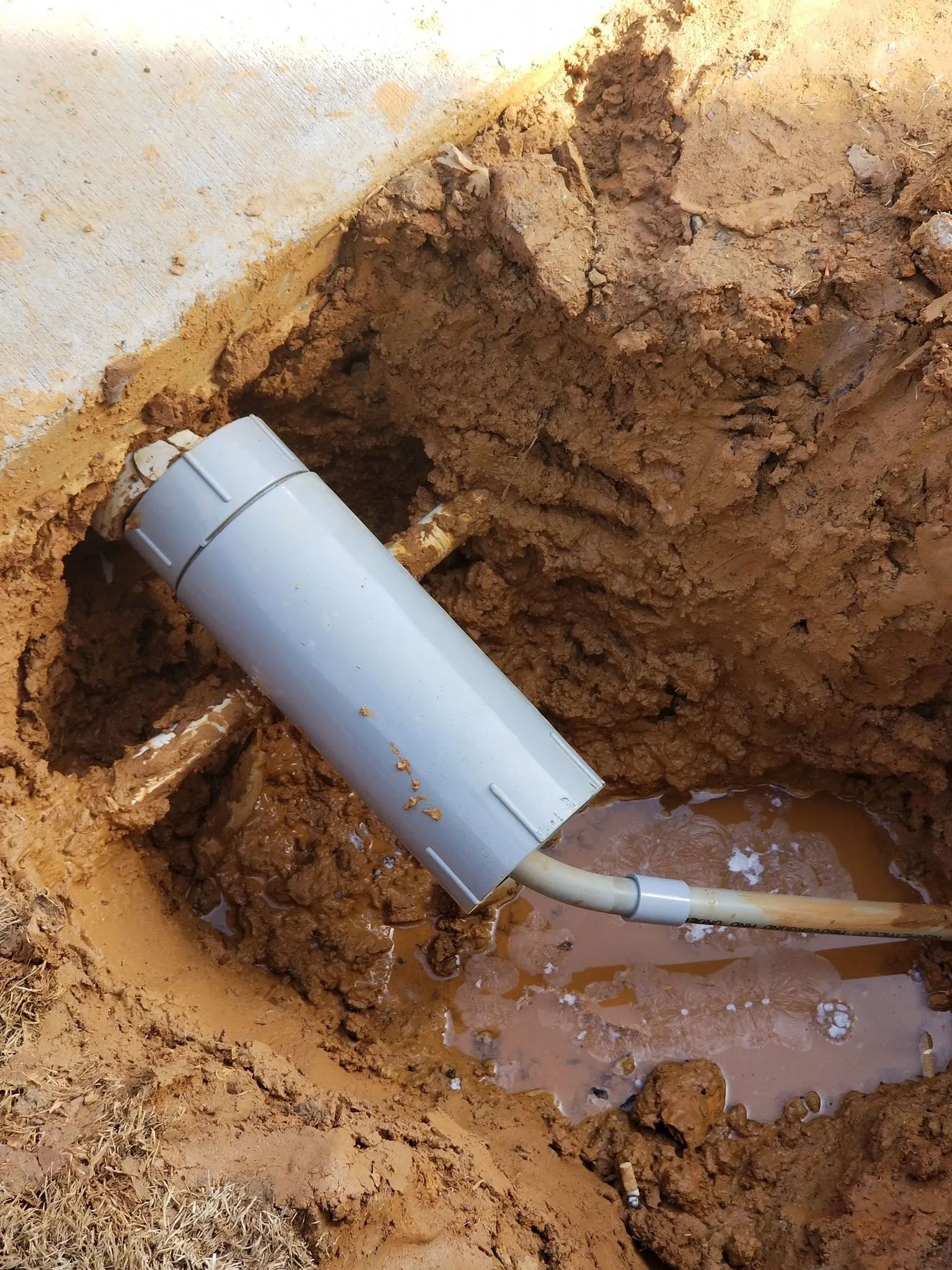 Broken gray pipe in muddy trench with muddy water pooling around it