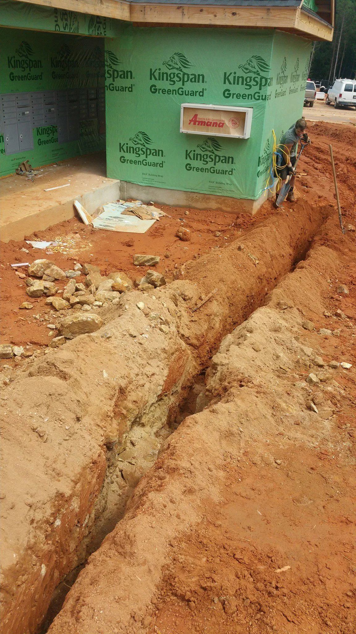 Dug trench beside a green building on a dirt roadside, with exposed earth and debris.