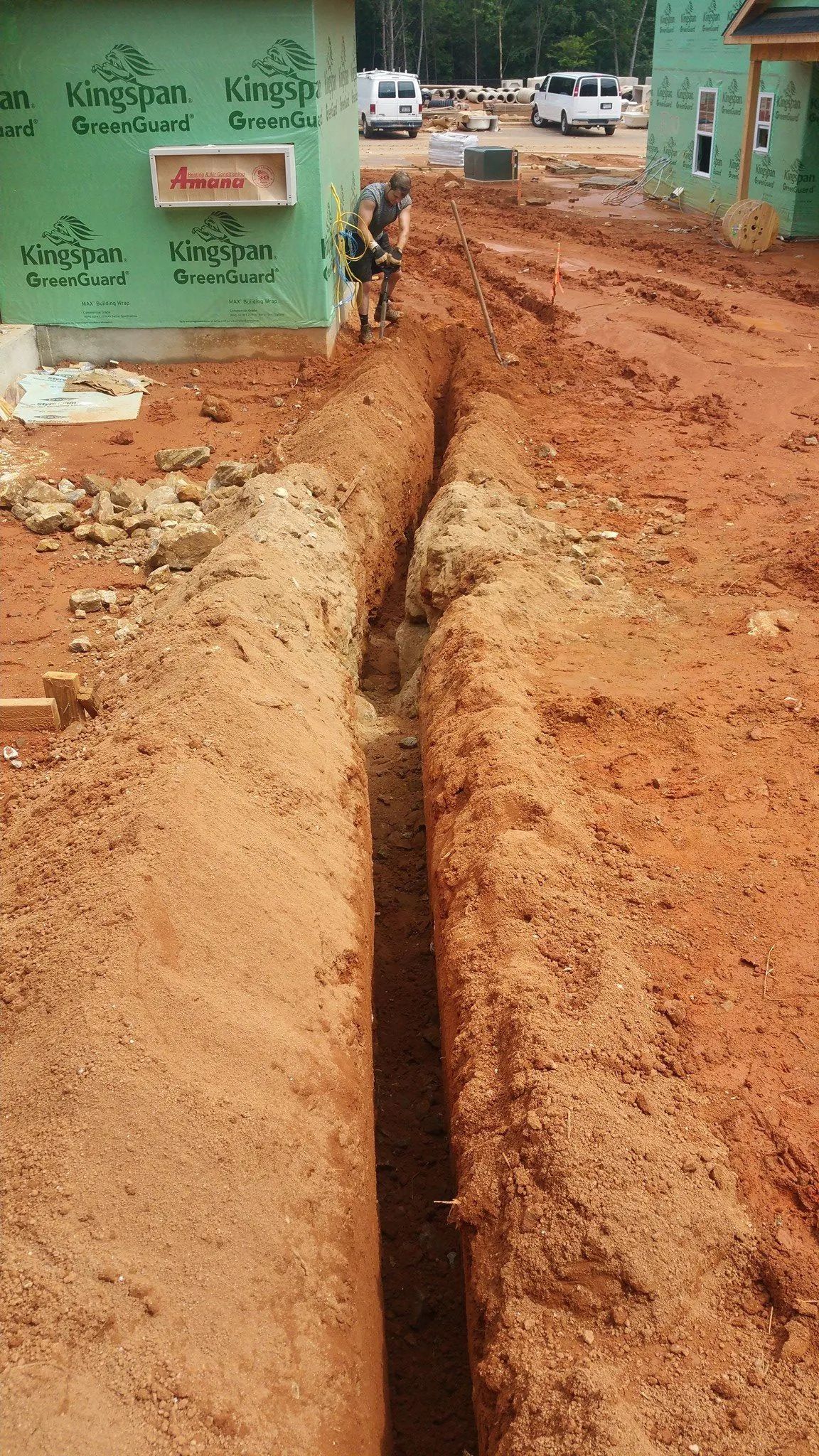 Dug trench along a red dirt street beside green buildings and parked vehicles, with a worker in the distance
