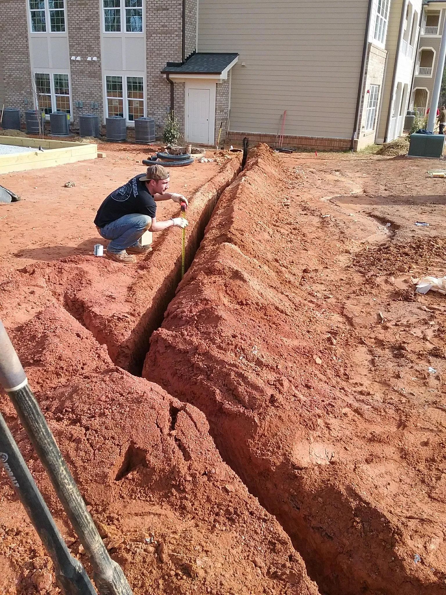 Worker crouching beside a deep trench in a red dirt construction site near apartment buildings