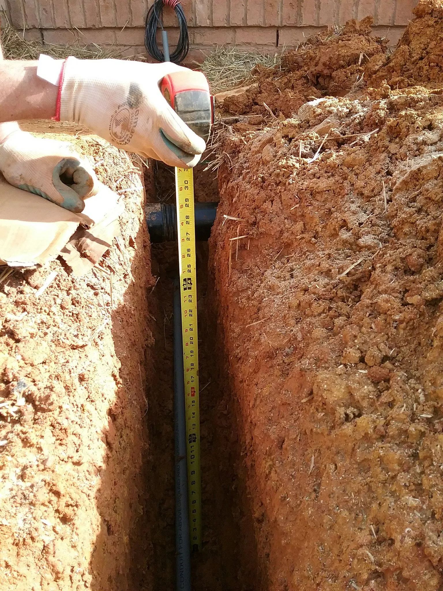 Gloved hands measuring the depth of a narrow trench with a tape measure and ruler.