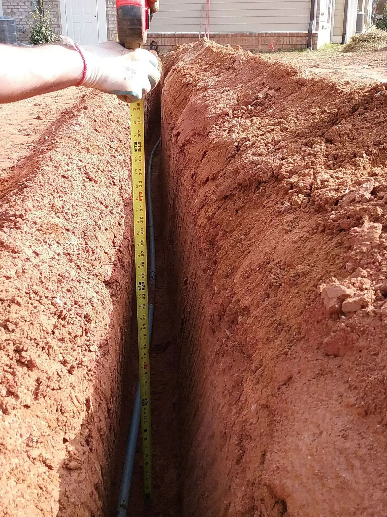 Deep trench with a measuring tape showing depth in red dirt outdoors