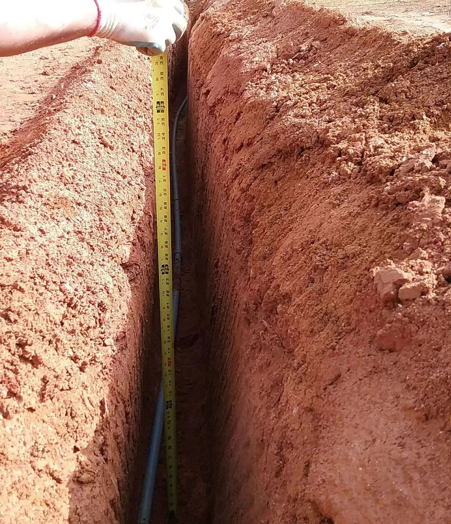 Hand holding a measuring tape in a narrow red dirt trench
