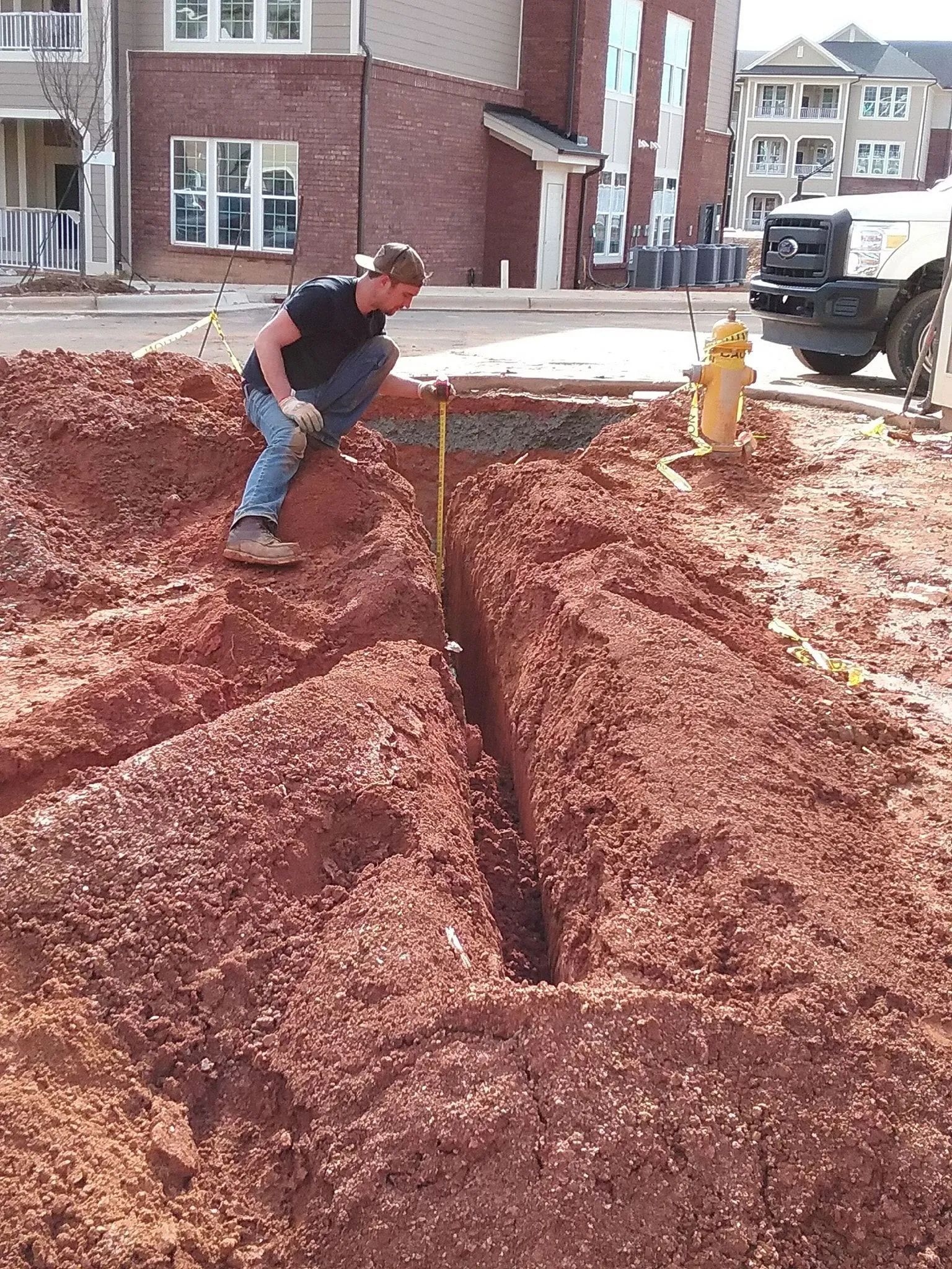 Worker in a trench beside a yellow fire hydrant at a construction site near brick houses.