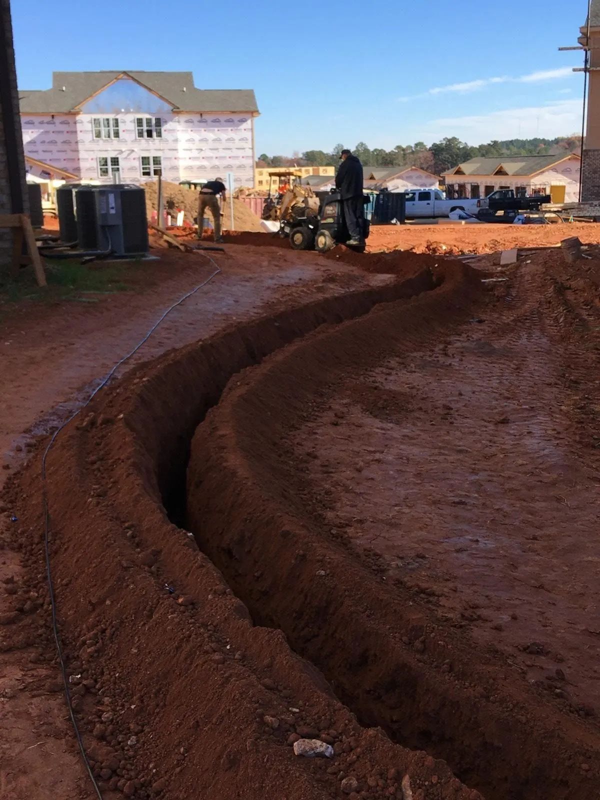 Curving dirt trench at a construction site with workers, equipment, and a white building in the background