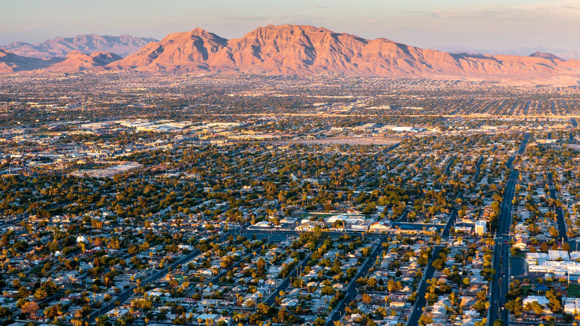 A sunlit aerial view of a sprawling residential neighborhood set against a backdrop of distant, rugged desert mountains.