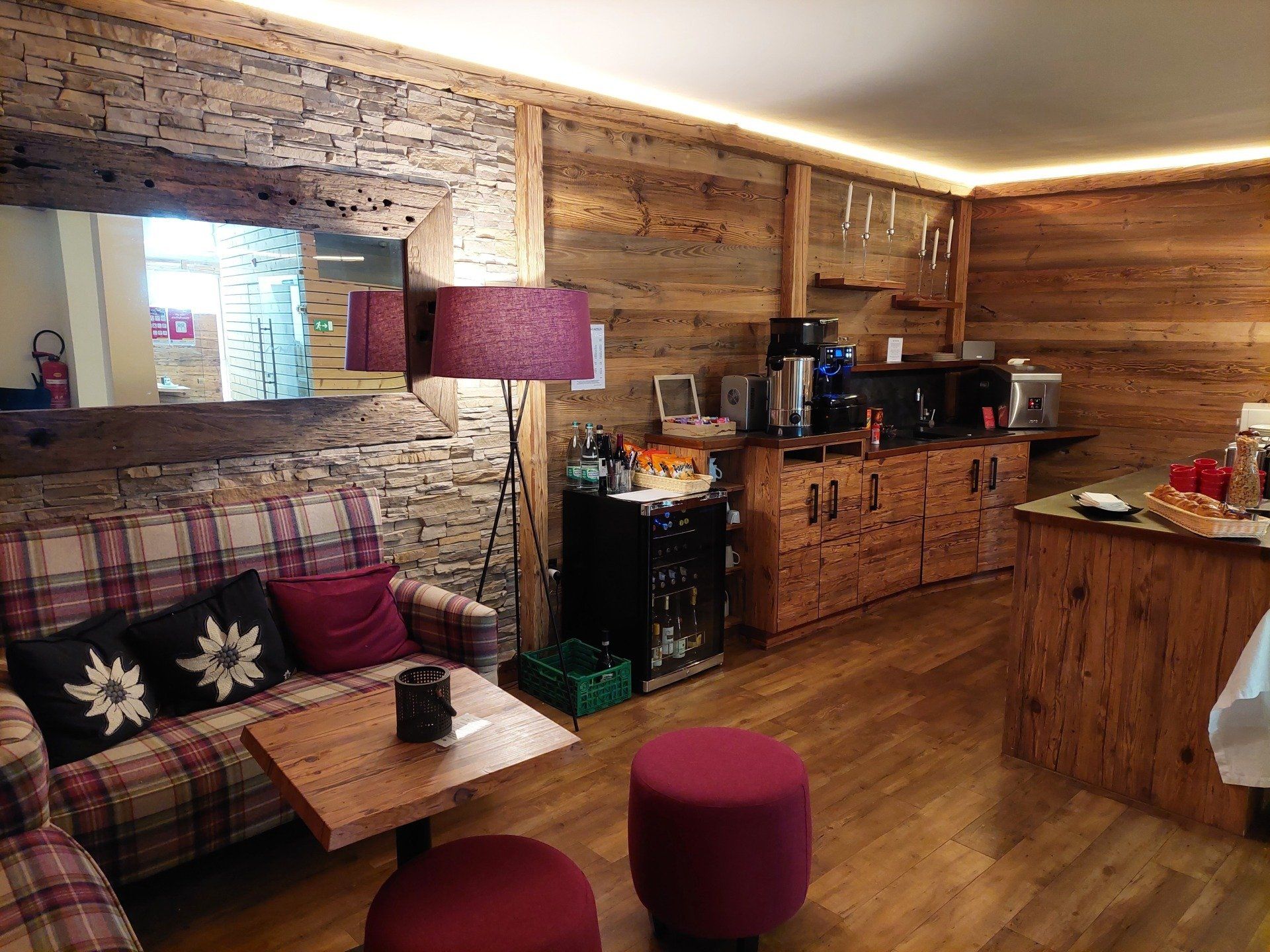 Cozy rustic living room with plaid sofa, magenta lamp and ottoman, wooden walls, and kitchen area in back