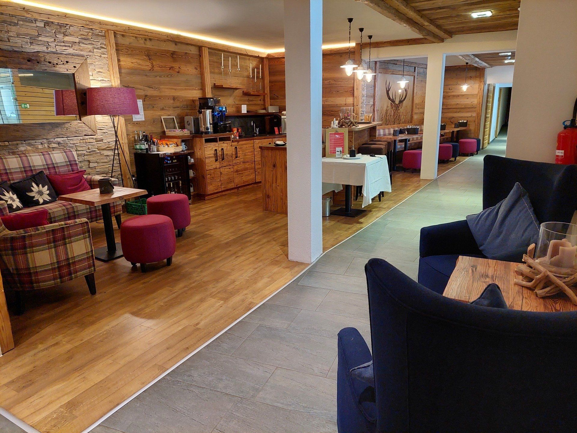 Cozy rustic lounge with wood paneling, plaid chairs, pink ottomans, and a dark blue sofa facing a dining area