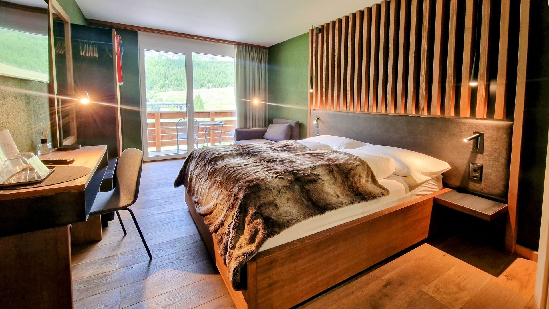 Modern hotel bedroom with a bed, fur throw, wooden slat headboard, and balcony doors overlooking greenery