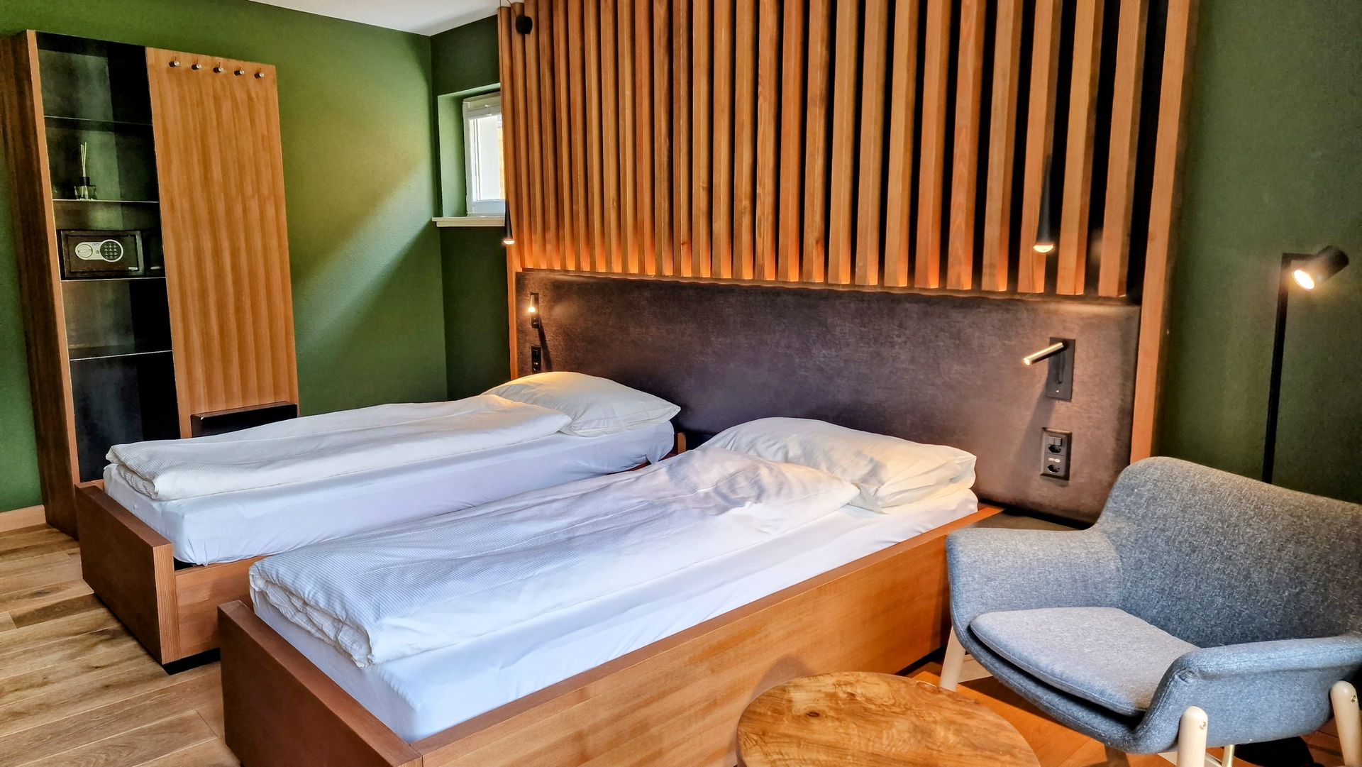 Modern hotel room with twin beds, wood slat headboard, green walls, and a gray armchair