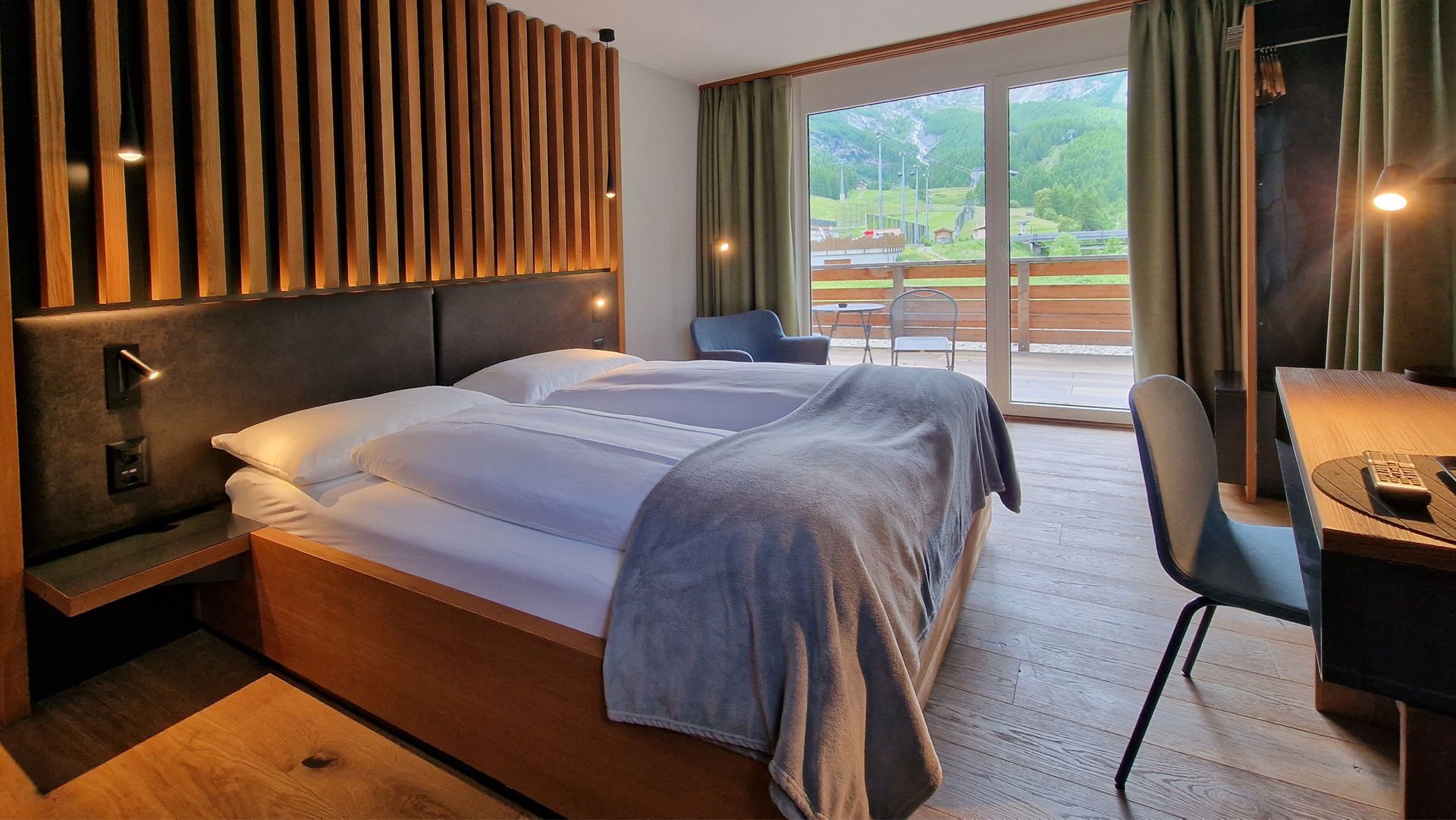 Modern hotel room with two beds, wood paneling, desk, and balcony doors overlooking a scenic view