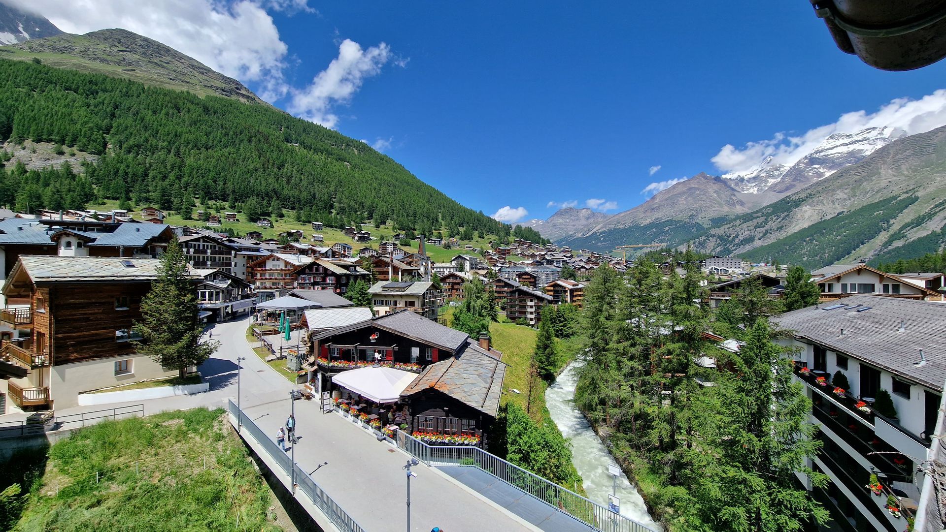 Mountain village with chalet-style buildings, green valley, and snow-capped peaks under a bright blue sky.