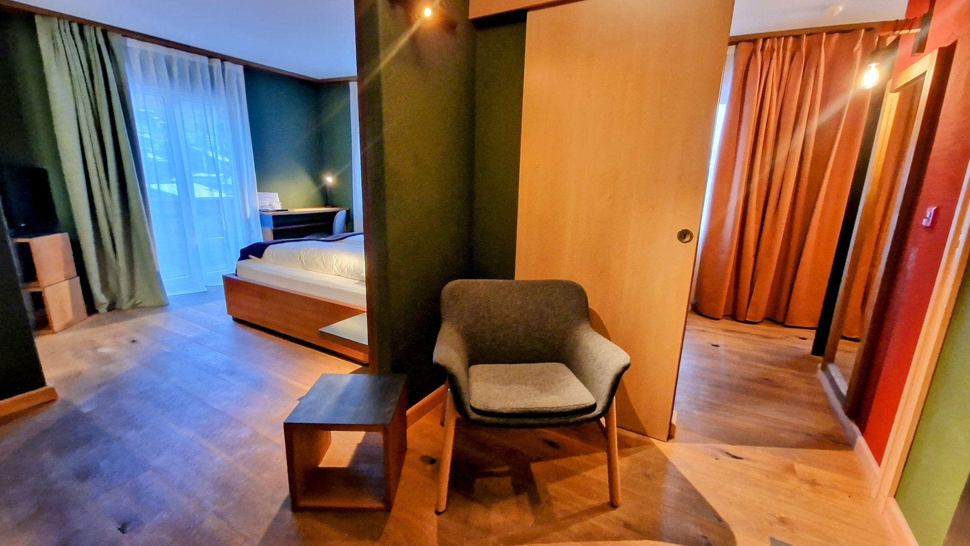Modern hotel room with a bed, chair, side table, and warm wood-paneled interior with curtains.