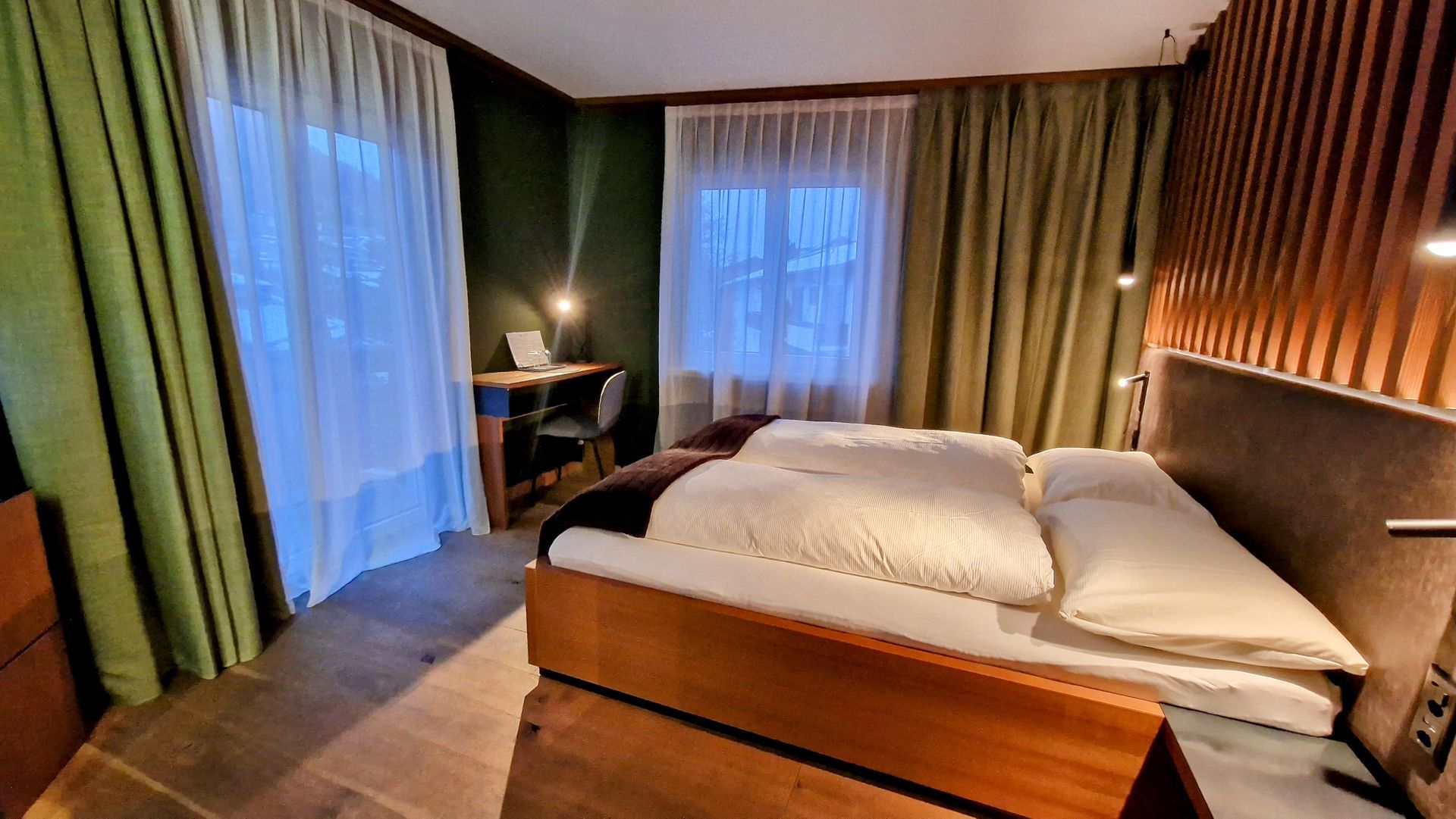 Modern hotel room with a made bed, curtains, wood accent wall, and balcony doors by the window