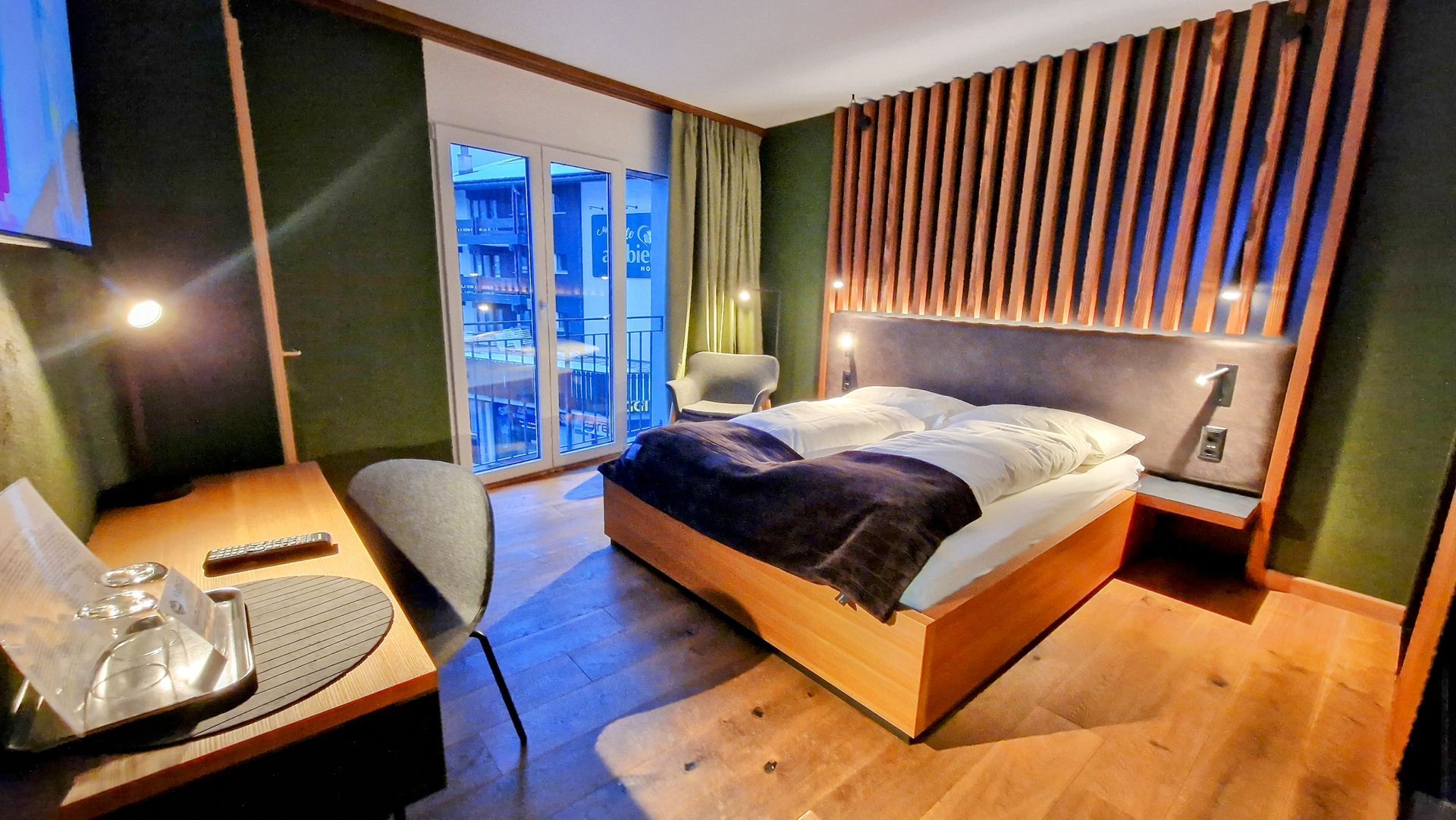 Cozy bedroom with a bed, wooden floor, desk, and blue-lit window view at dusk