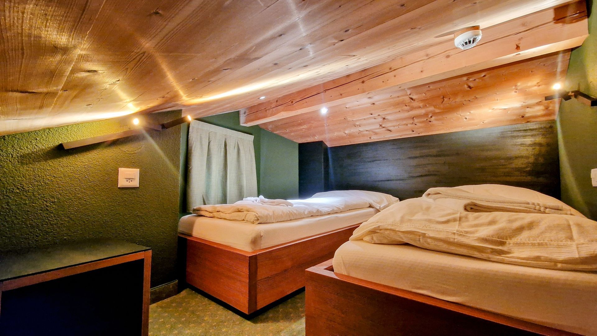 Cozy attic bedroom with two beds, green walls, wood-paneled ceiling, and warm lighting