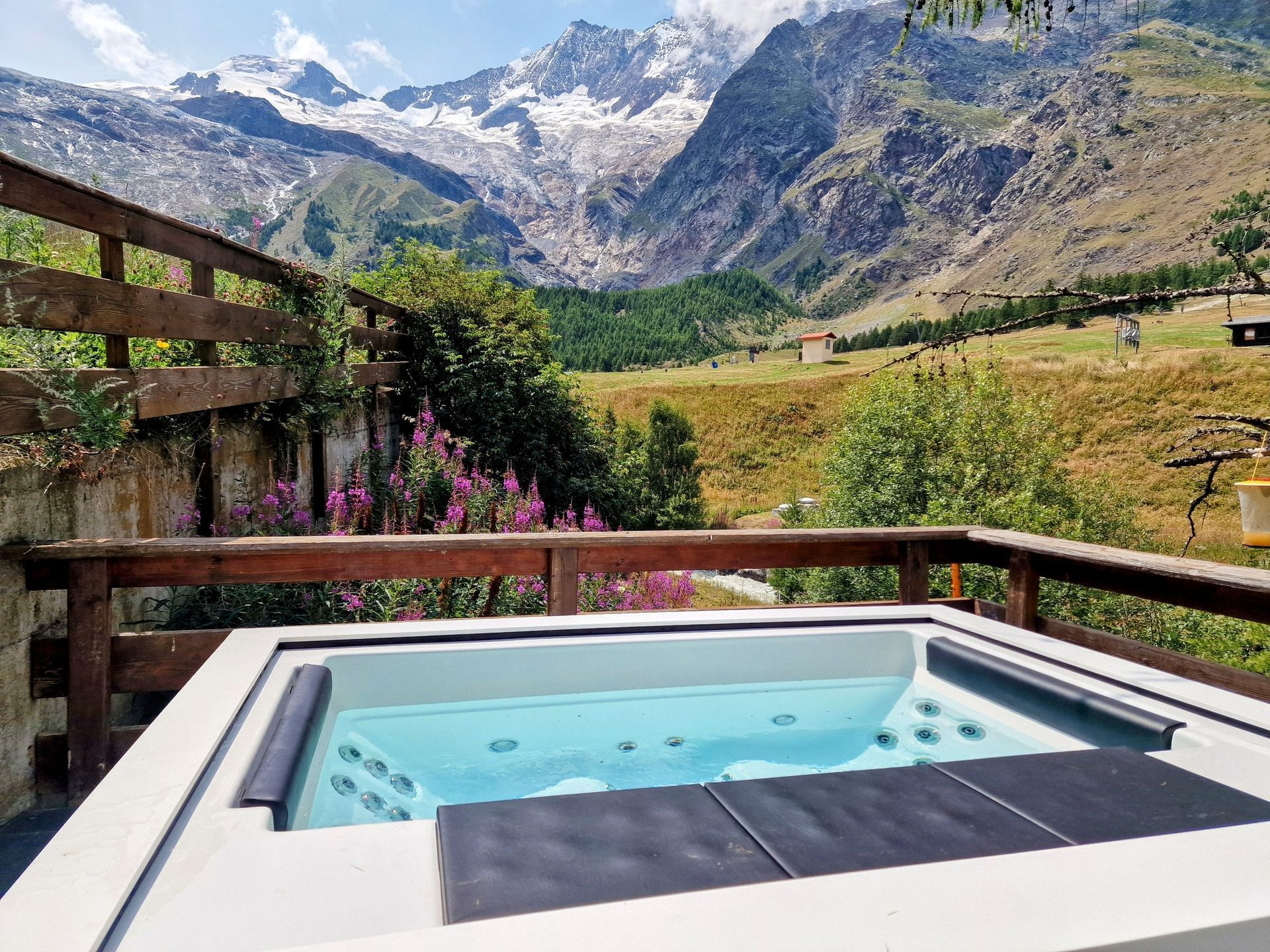 Hot tub on a wooden deck overlooking alpine mountains and a grassy valley with wildflowers