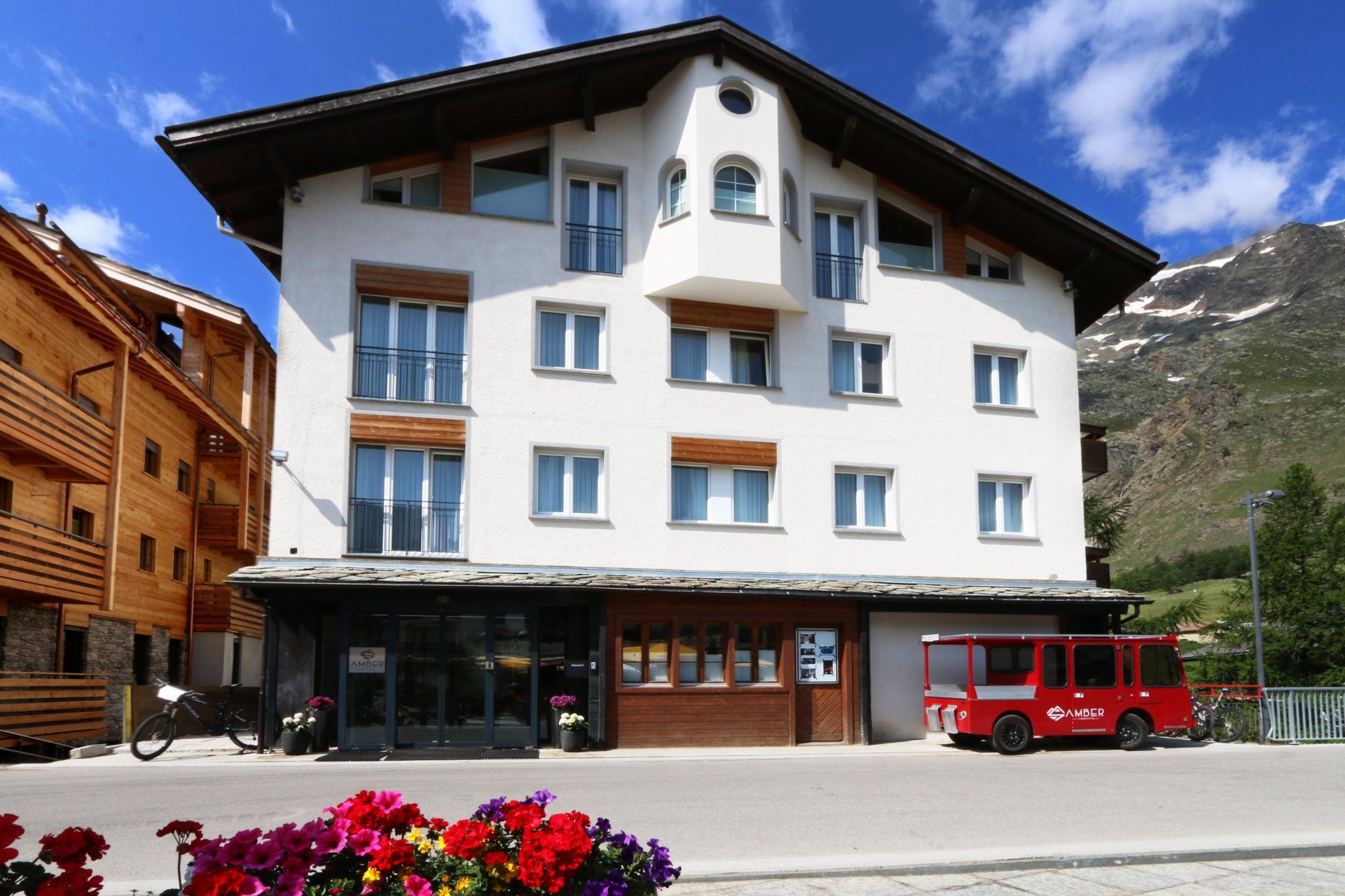 White mountain hotel with balconies and a red van parked outside under a blue sky.