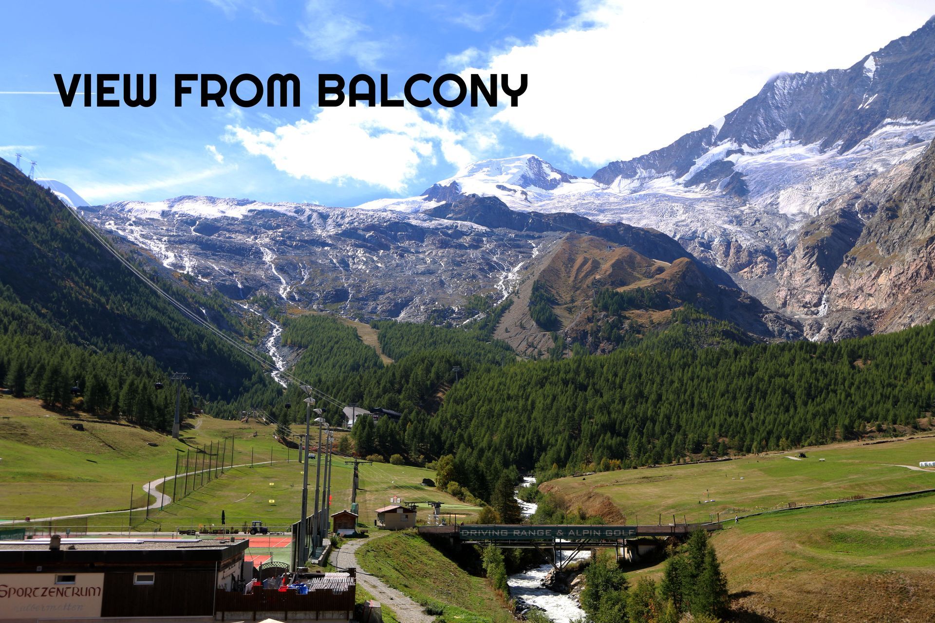 Mountain valley view from a balcony with green slopes, forest, and snow-capped peaks under a bright sky