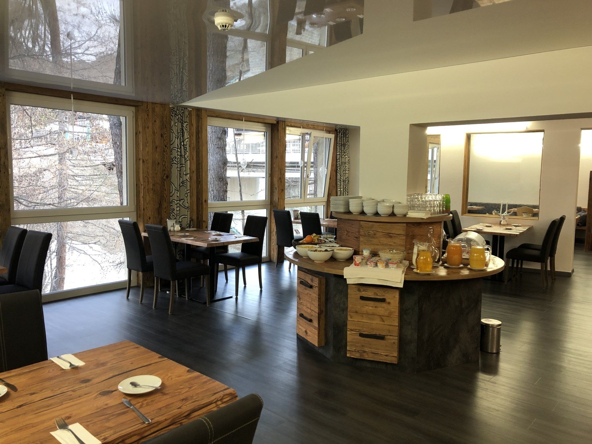 Modern hotel lounge with large windows, dark floors, wooden tables, and a central breakfast buffet area