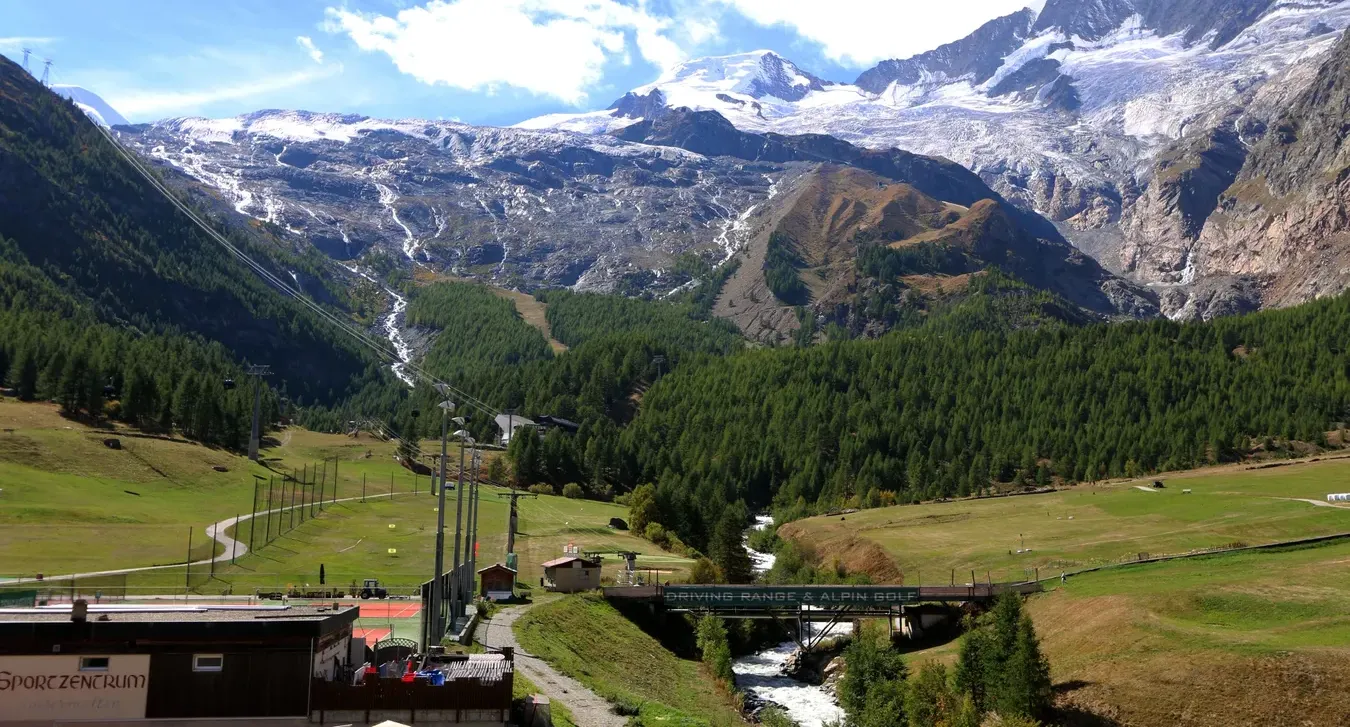 Mountain valley with green meadows, pine forests, snowy peaks, and a small bridge under a bright blue sky