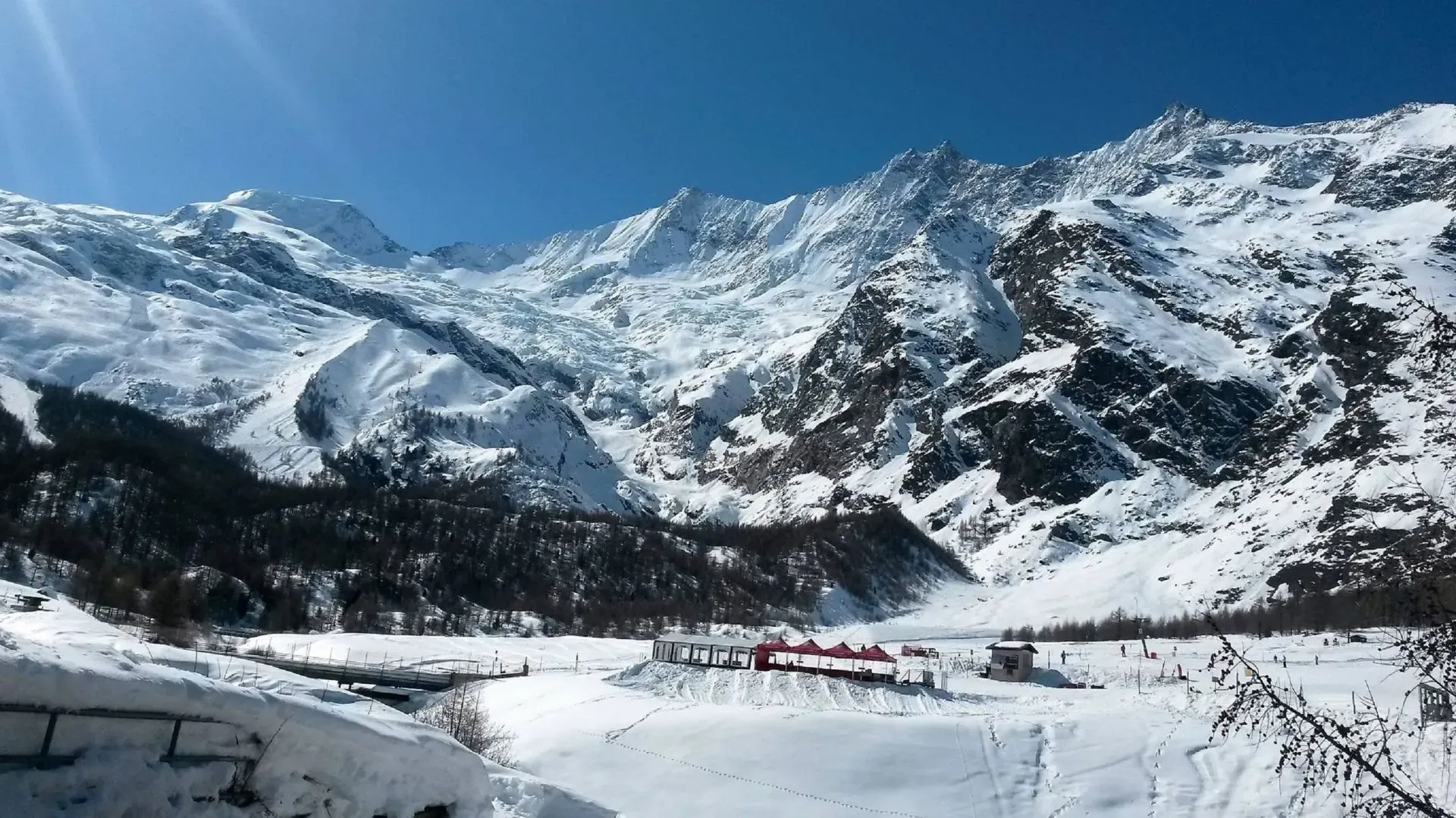 Snow-covered mountain valley with a frozen river and small red buildings under a clear blue sky