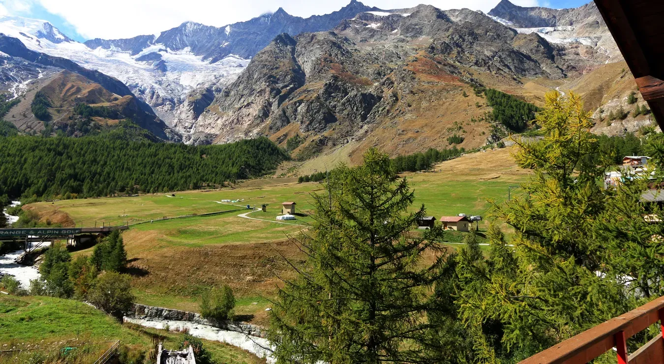 Mountain valley with snow-capped peaks, green meadow, and a small stream under a clear sky
