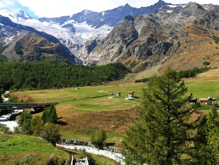 Mountain valley with green meadow, winding stream, pine trees, and snow-capped peaks under a clear sky