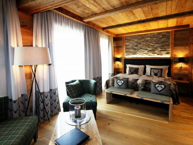 Cozy rustic bedroom with wooden beams, a large bed, armchair, lamp, and floor-to-ceiling curtains