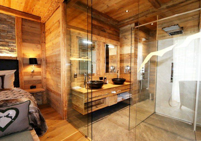 Wood-paneled bathroom with glass shower, double sink vanity, and a bedroom visible through the open doorway